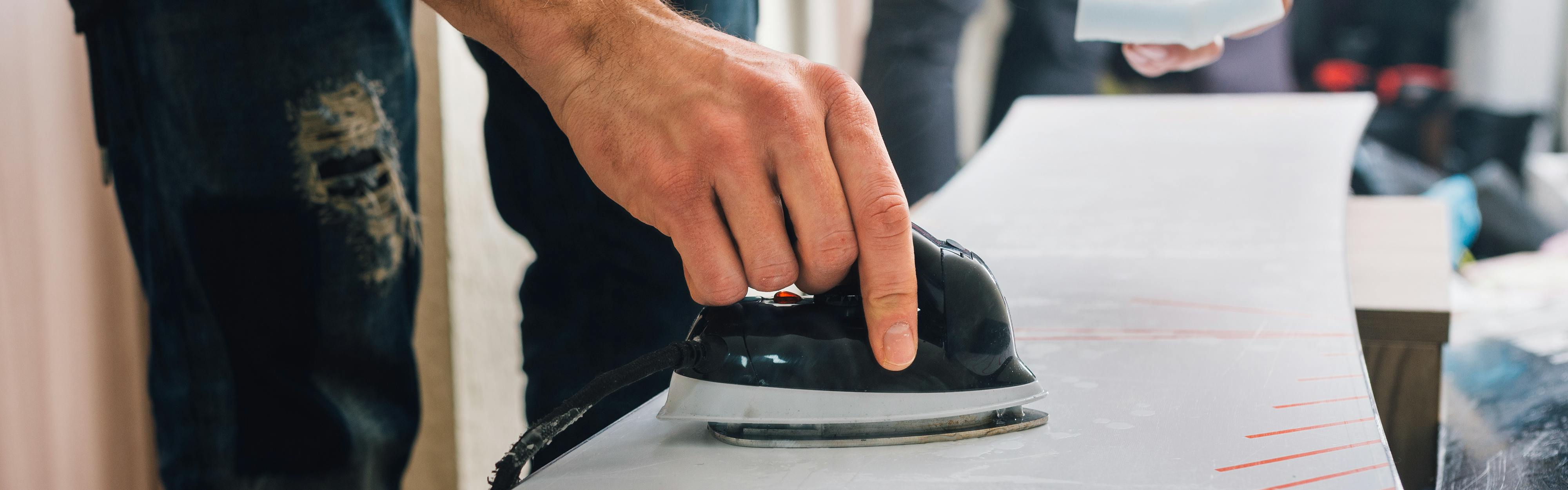 A man waxing a snowboard with a small iron. 