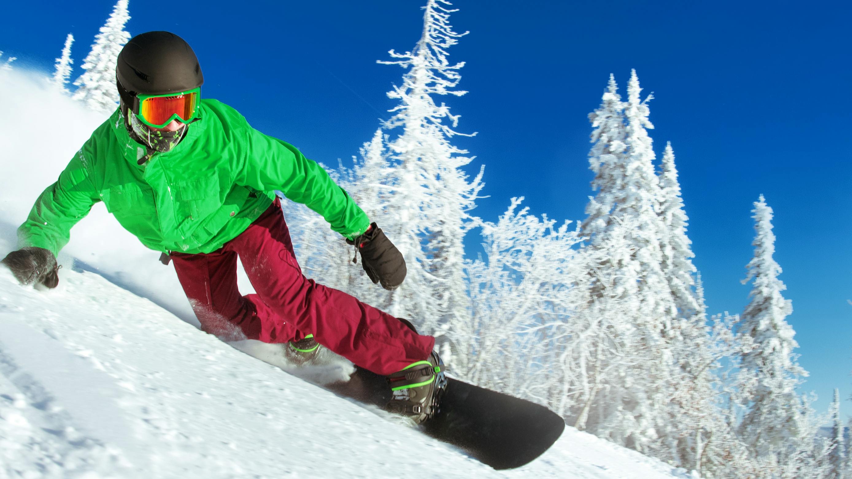 A snowboarder carving down a ski run in red pants and a green jacket. 