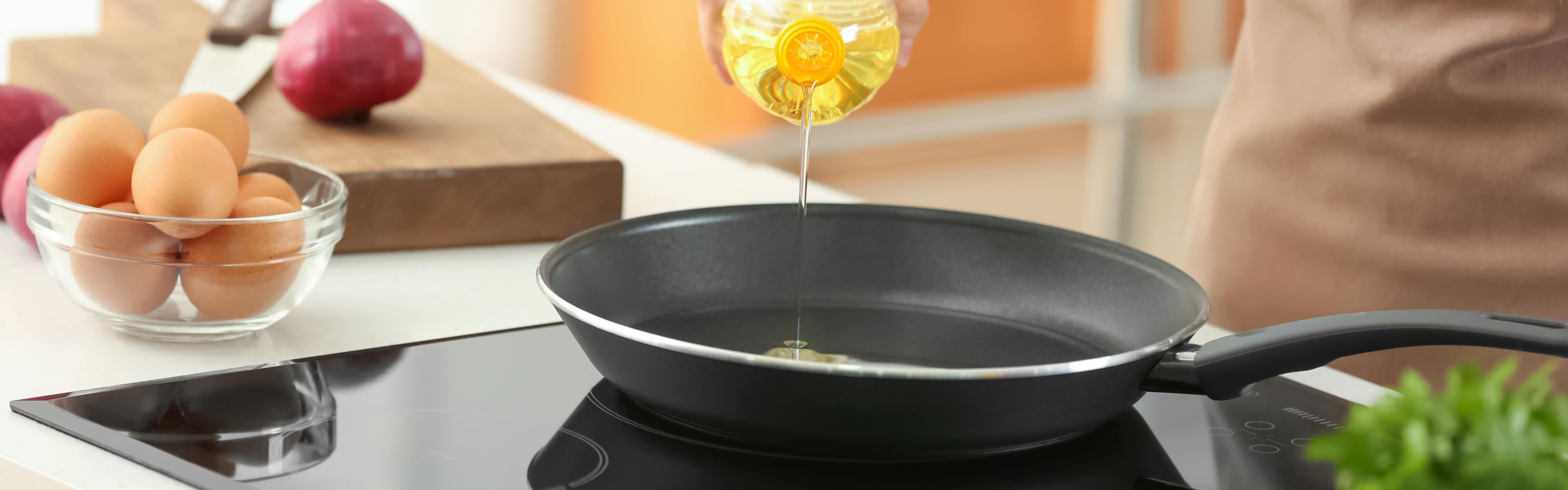 A hand pouring oil from a bottle into a pan on an induction stove. There is a bowl of eggs sitting next to the stove. 
