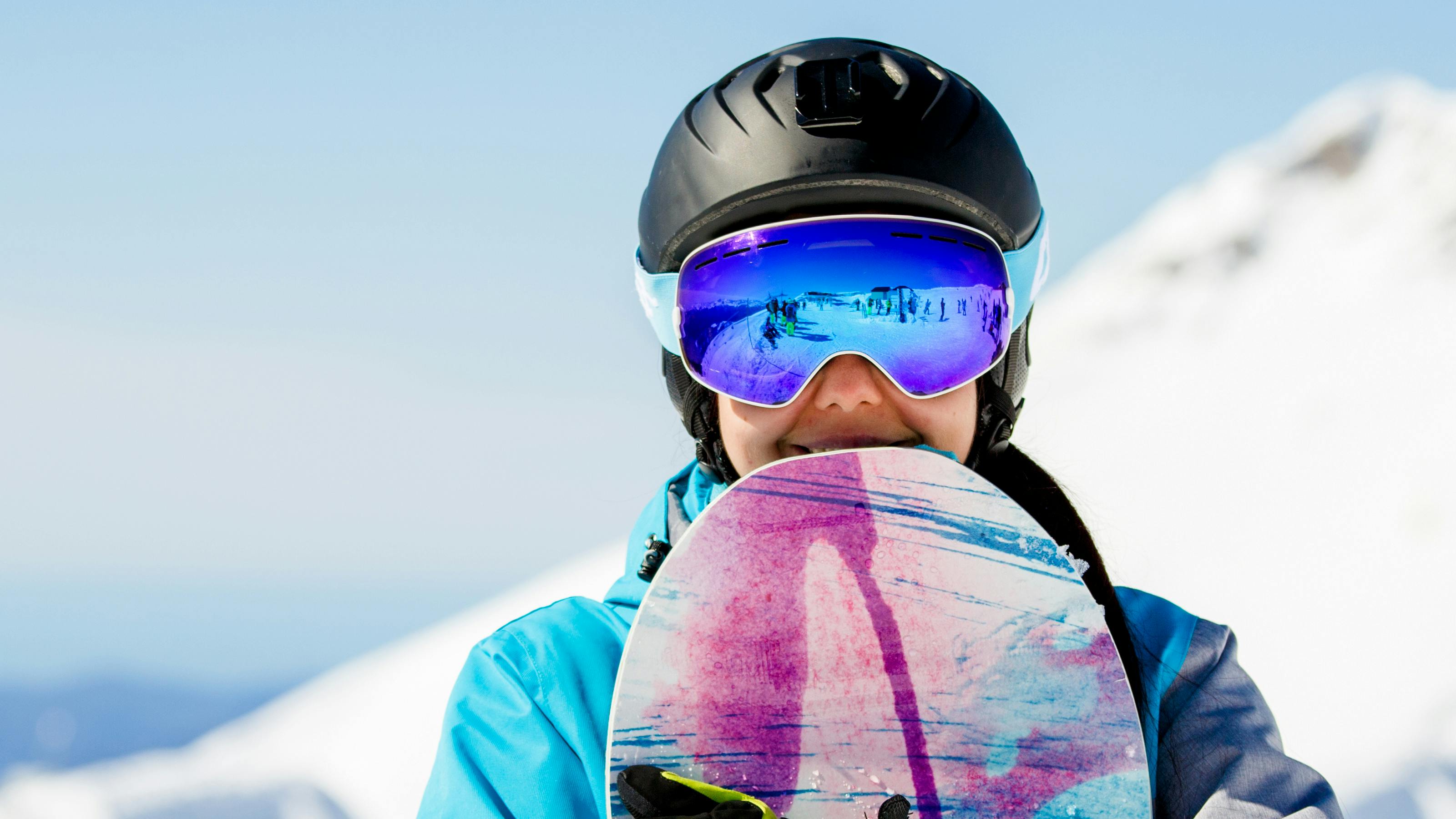 A snowboarder holding her board as she looks at the camera. She is wearing goggles and a helmet. 
