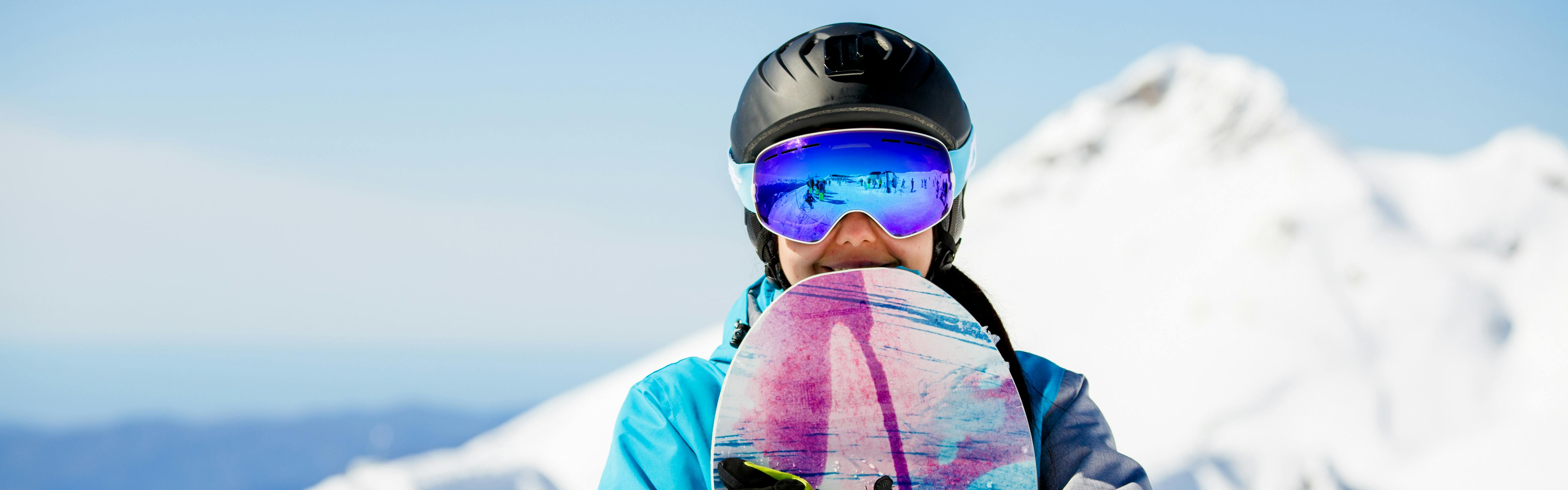 A snowboarder holding her board as she looks at the camera. She is wearing goggles and a helmet. 