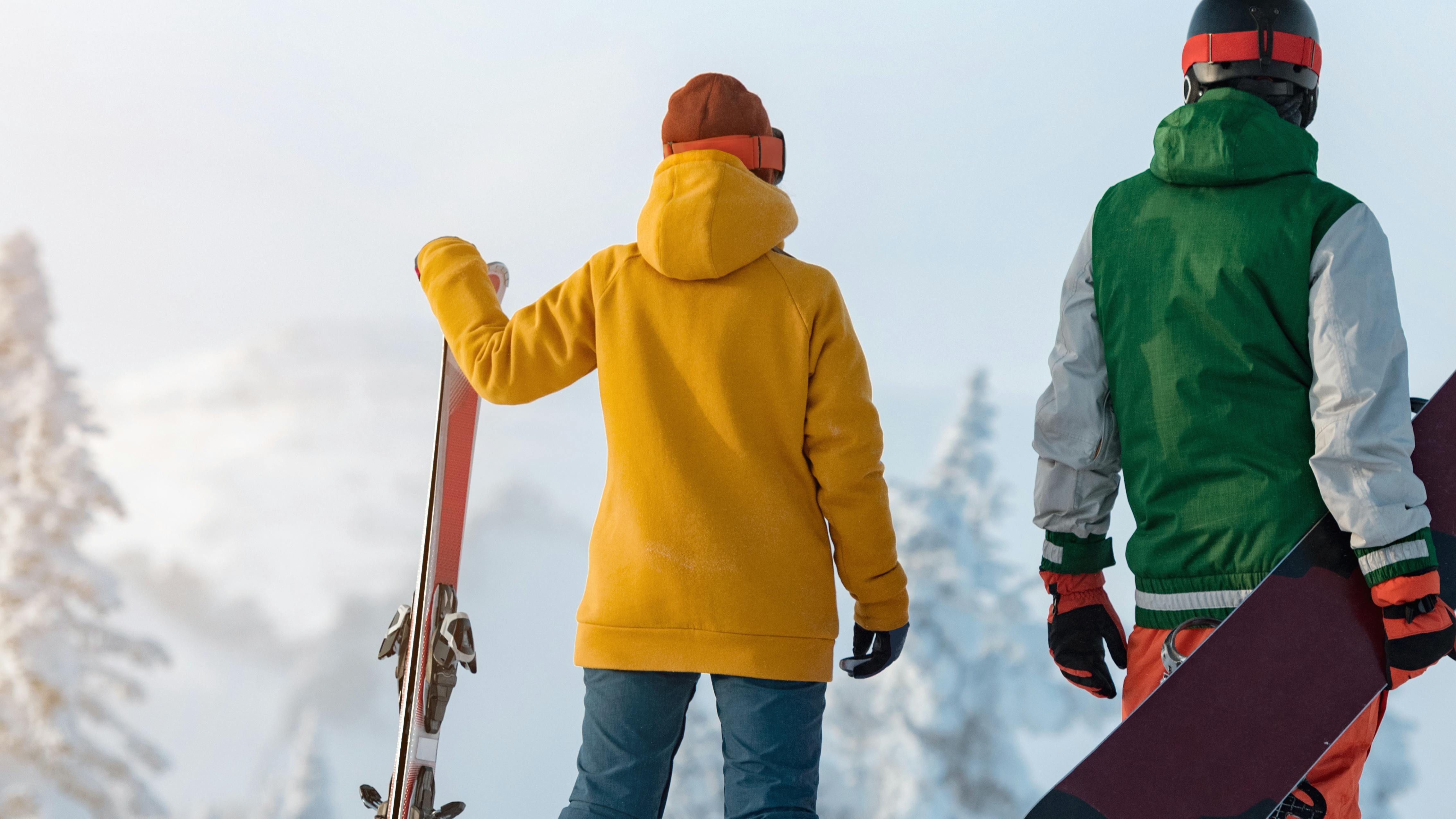 A skier and snowboarder standing at the top of a ski run holding their gear. 