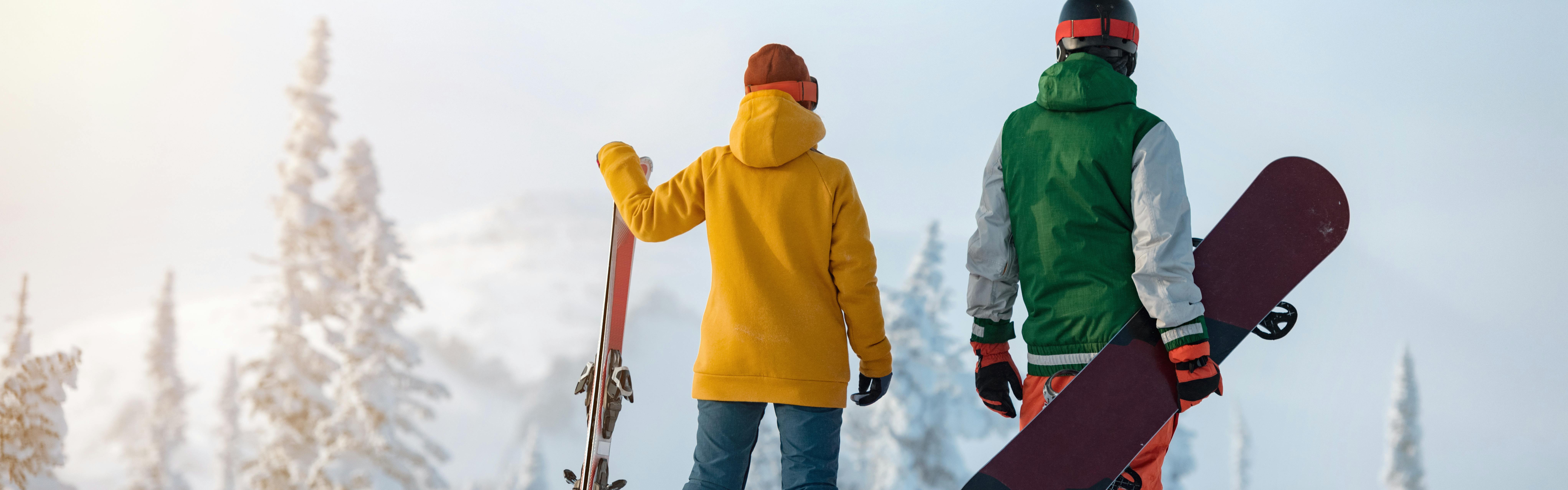A skier and snowboarder standing at the top of a ski run holding their gear. 