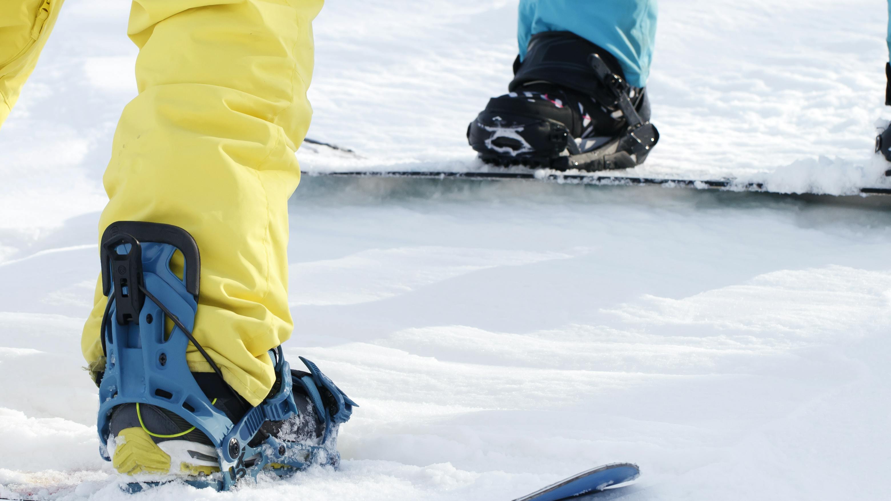 Two snowboarders stand in their snowboard boots and bindings in the snow. The image is cropped so just their boots, bindings, and boards are visible. 