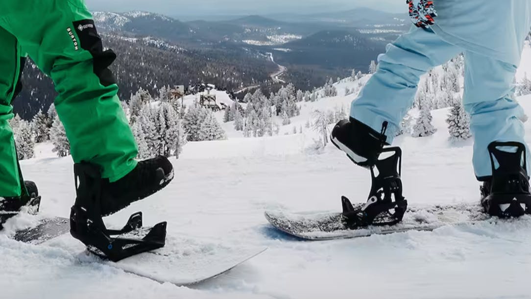 Two snowboarders standing on the top of a snowy mountain as they put their boots in their bindings. 