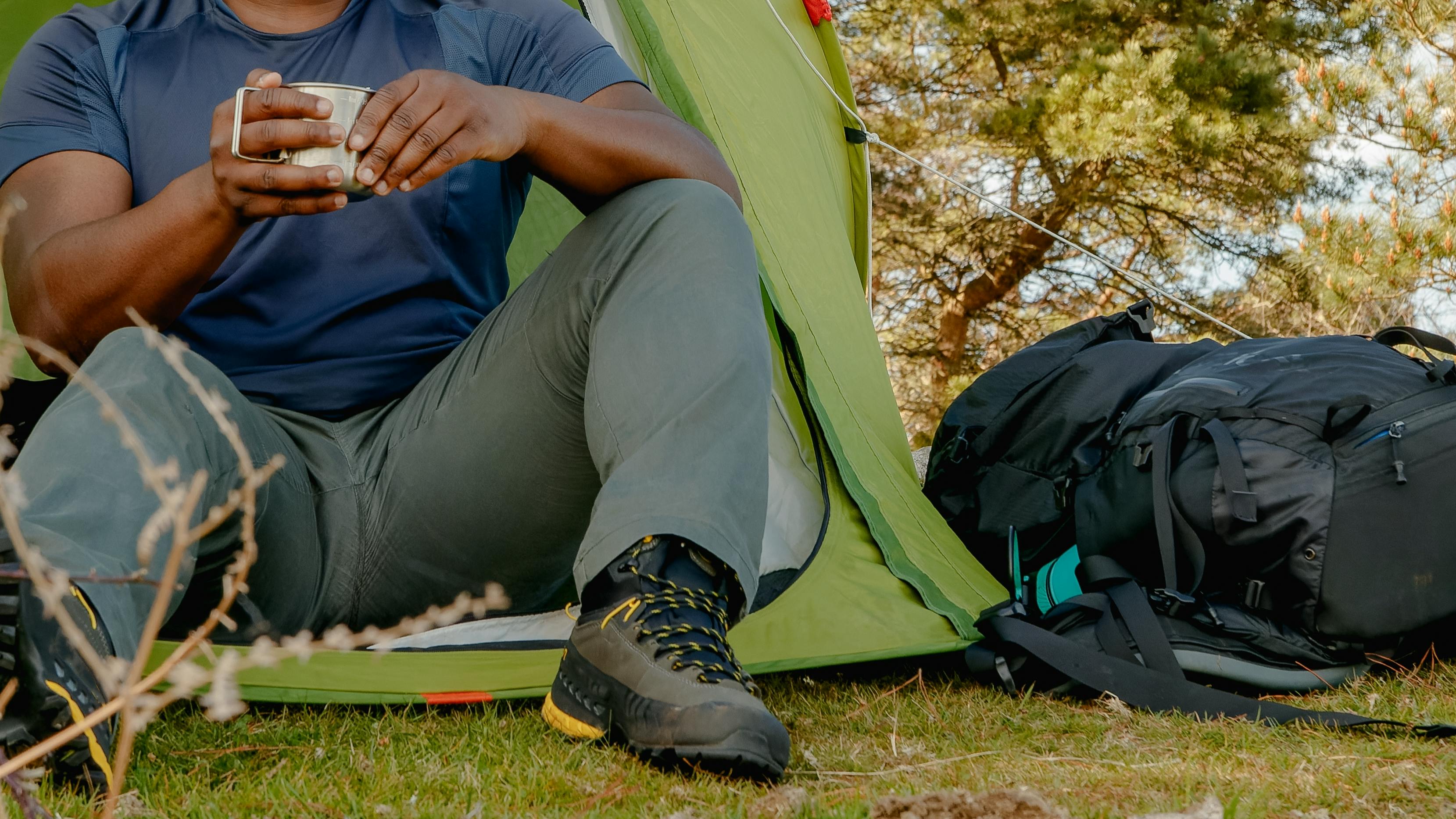 A hiker sitting in his tent as he holds a coffee cup. There is a backpack sitting next to his tent. 