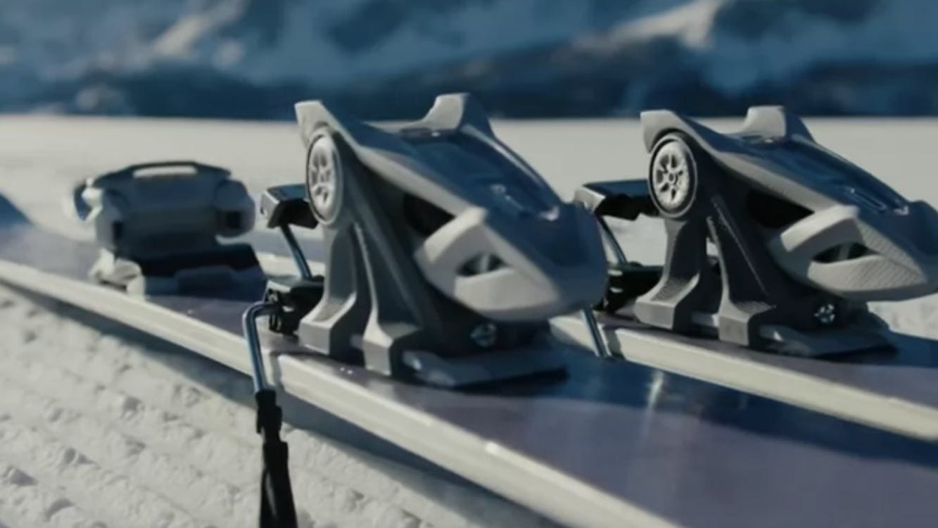 Marker ski bindings mounted to skis sitting on a snowy surface. There are mountains in the background. 