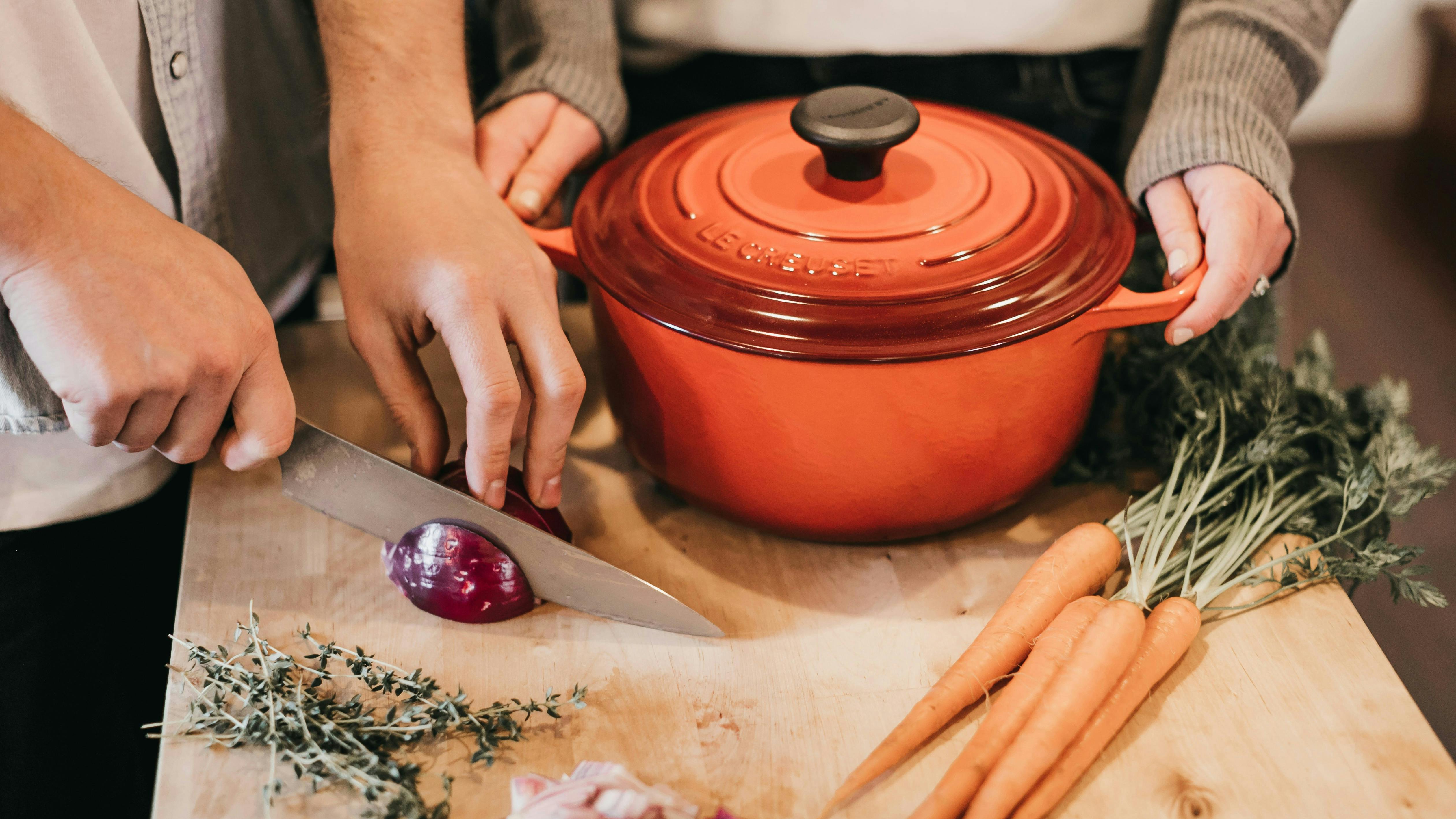 A red Le Creuset Dutch Oven on a wooden cutting board as one person holds the handles and another person cuts an onion on the same wood cutting board. There are some carrots and some thyme on the cutting board. 
