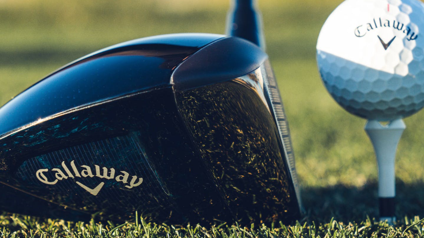 Close up of a Callaway driver in front of a Callaway golf ball. There are blurred feet in the background and the driver is resting on the grass. 