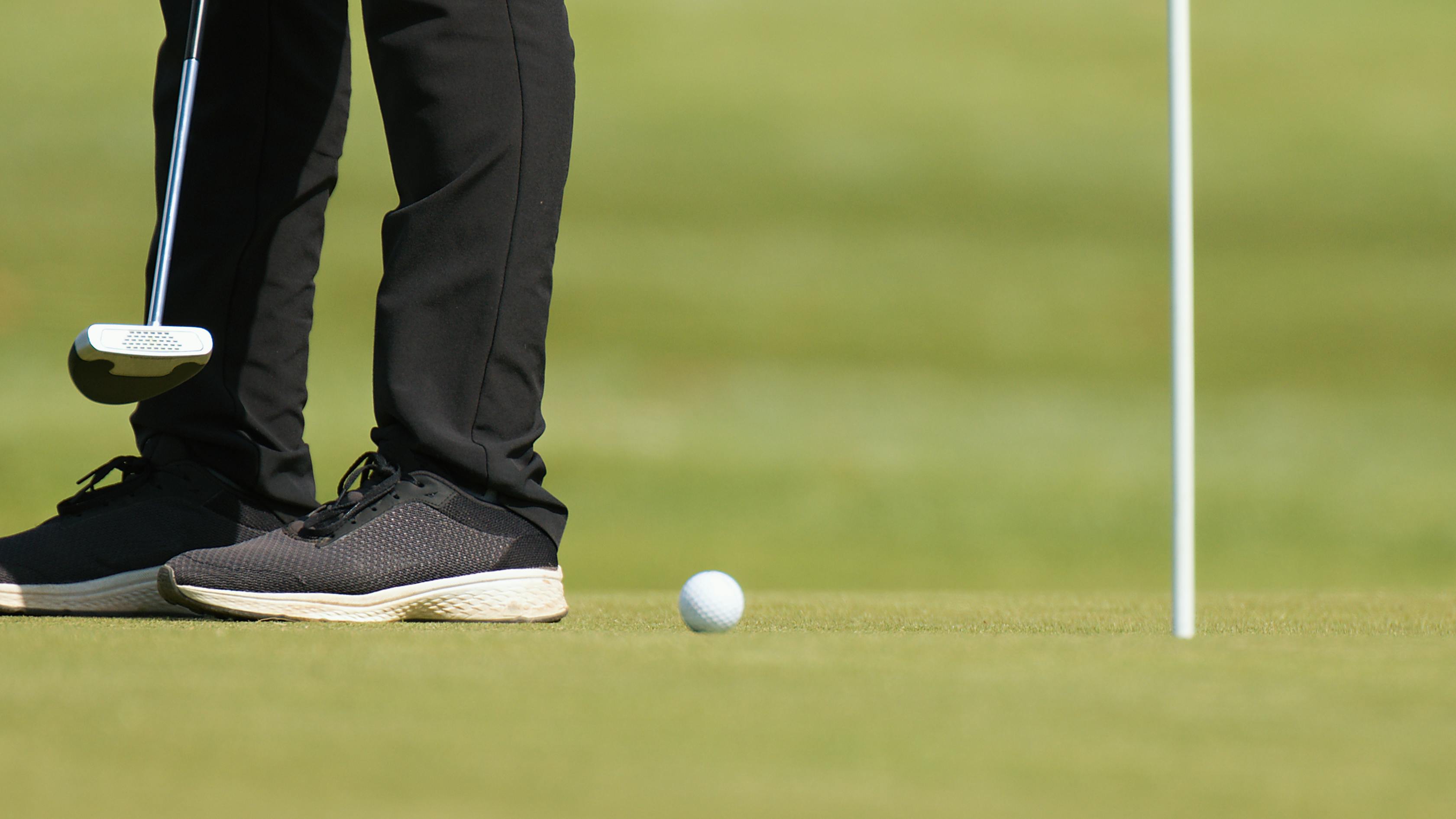 Close up of a golfers shoes as hes hitting a golf ball with a putter. 