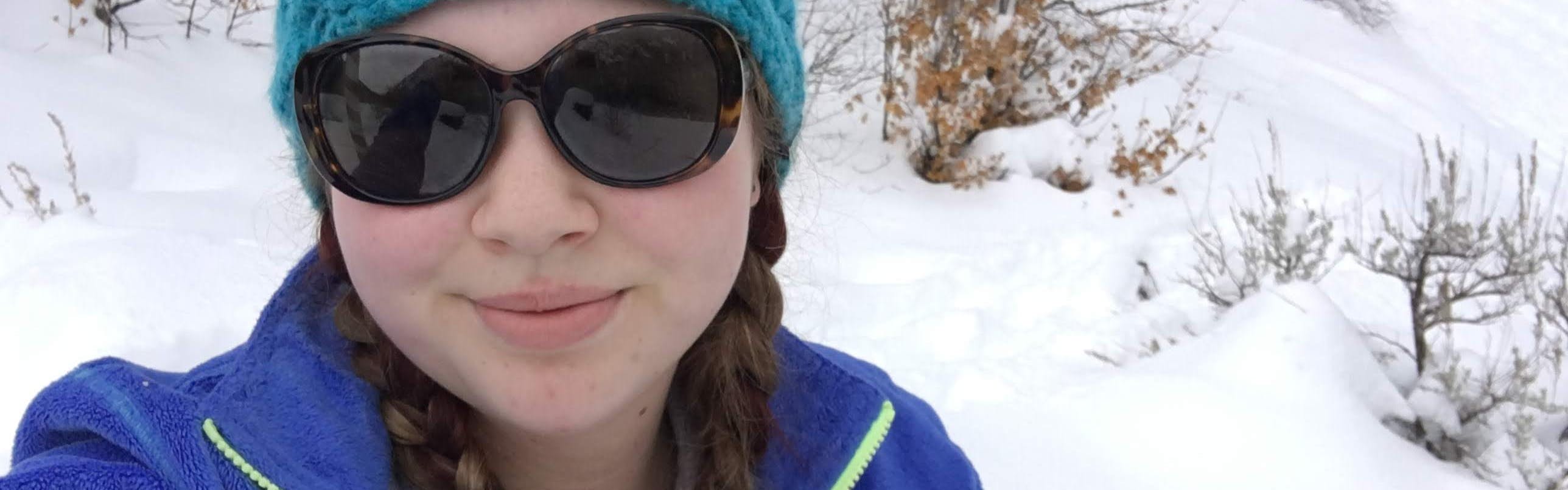 Young woman is sitting in front of a snowbank. She is wearing a blue knitted hat, large sunglasses and a blue fleece jacket. Her hair is in two braids. 