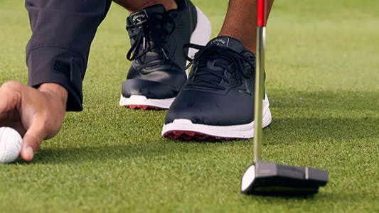 A golfer setting a golf ball on the green. He is wearing Callaway golf shoes and is holding a Callaway club. 