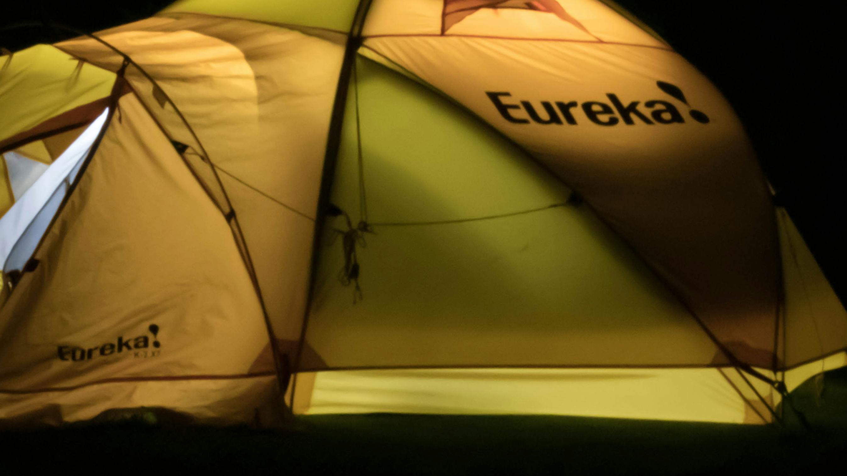 A Eureka tent lit up from inside as it's dark outside. The tent is green. 
