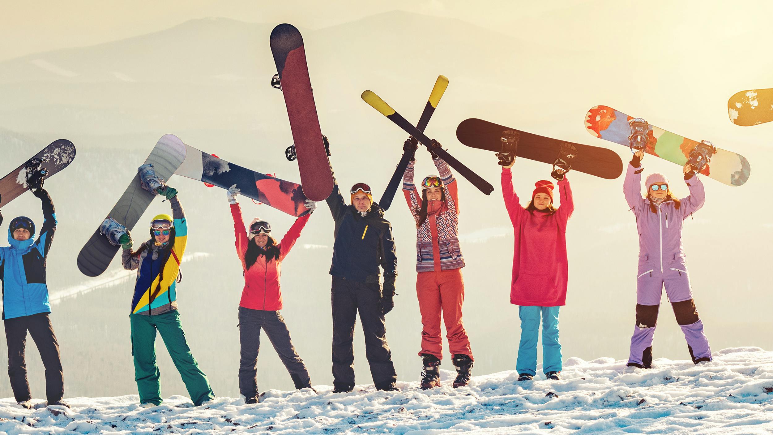 Several snowboarders wearing different outfits stand at the top of a snowy mountain while holding their snowboards in the air. 