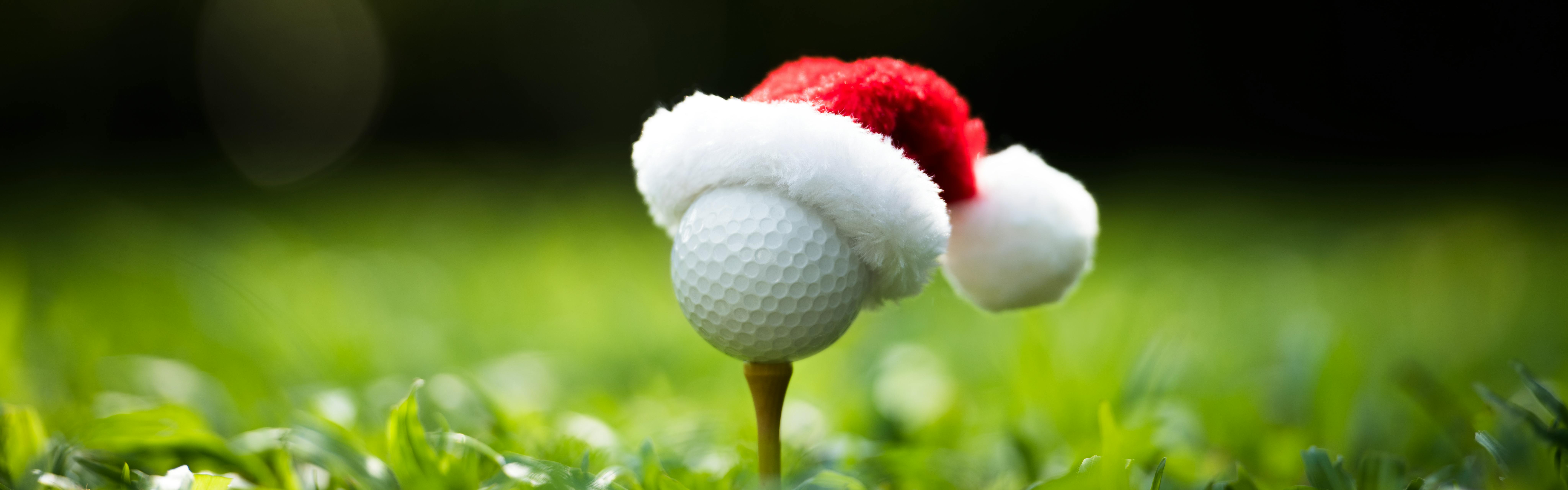 A golf ball on a tee wearing a small red and white santa hat. 