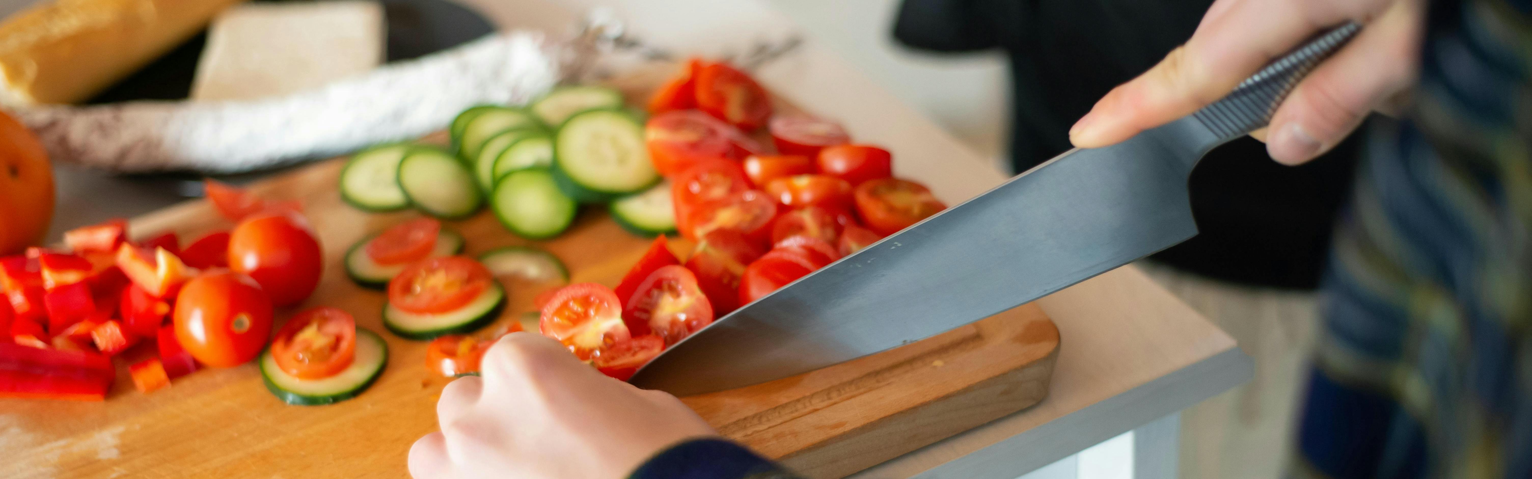 An at-home chef uses a knife to slice tomatoes and cucumber on a cutting board.