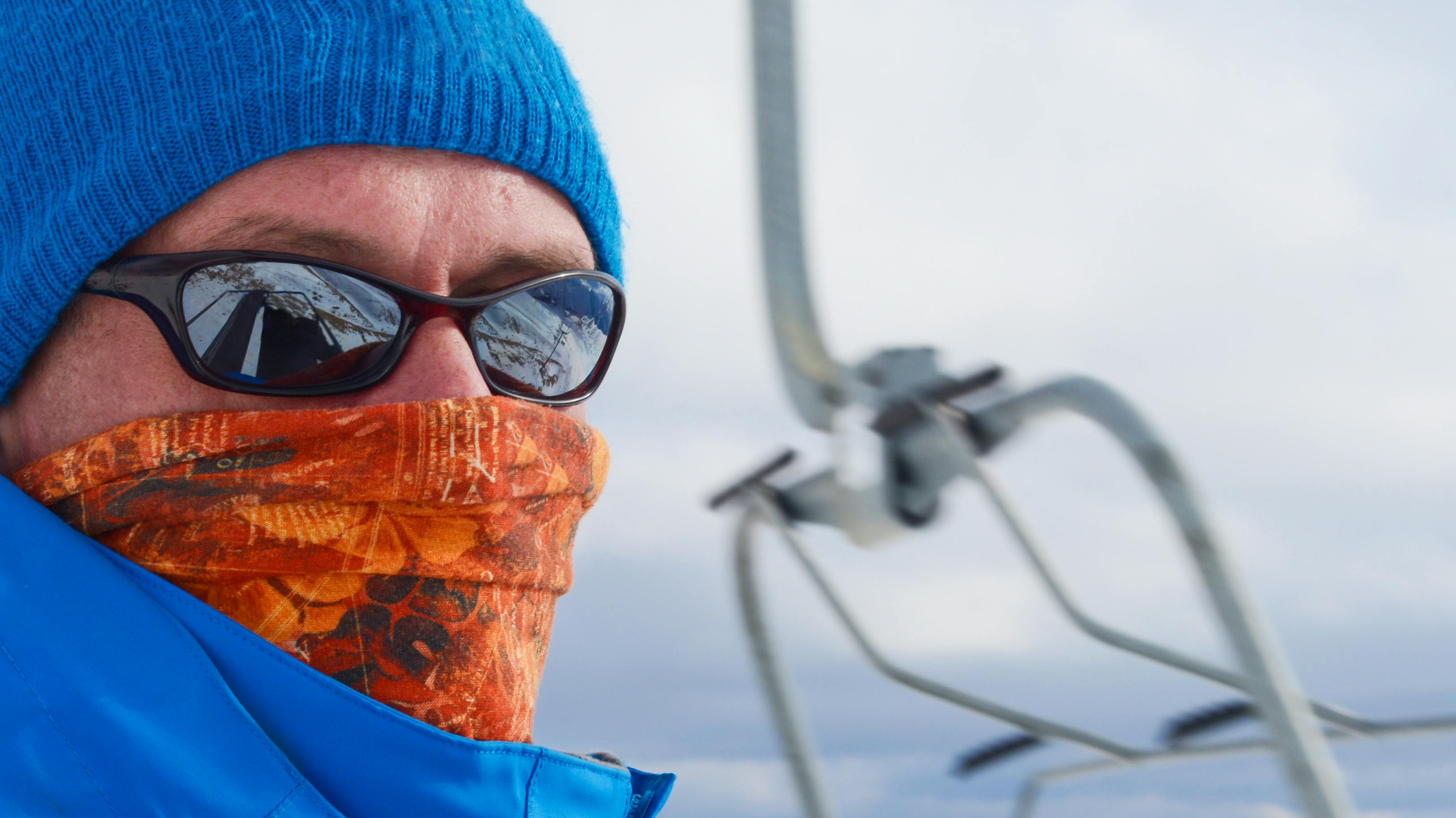 A snowboarder sits on the chairlift with a facemask on and sunglasses covering his eyes. 
