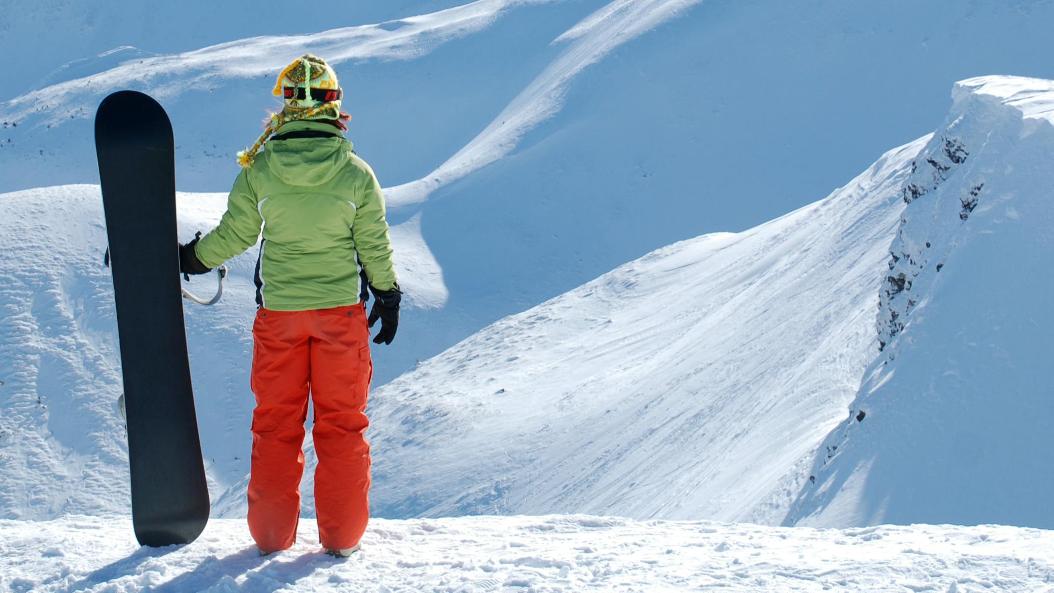 A woman with a snowboard stands at the top of a snowy mountain. Her back is facing the camera and she has a green jacket and red pants on. 