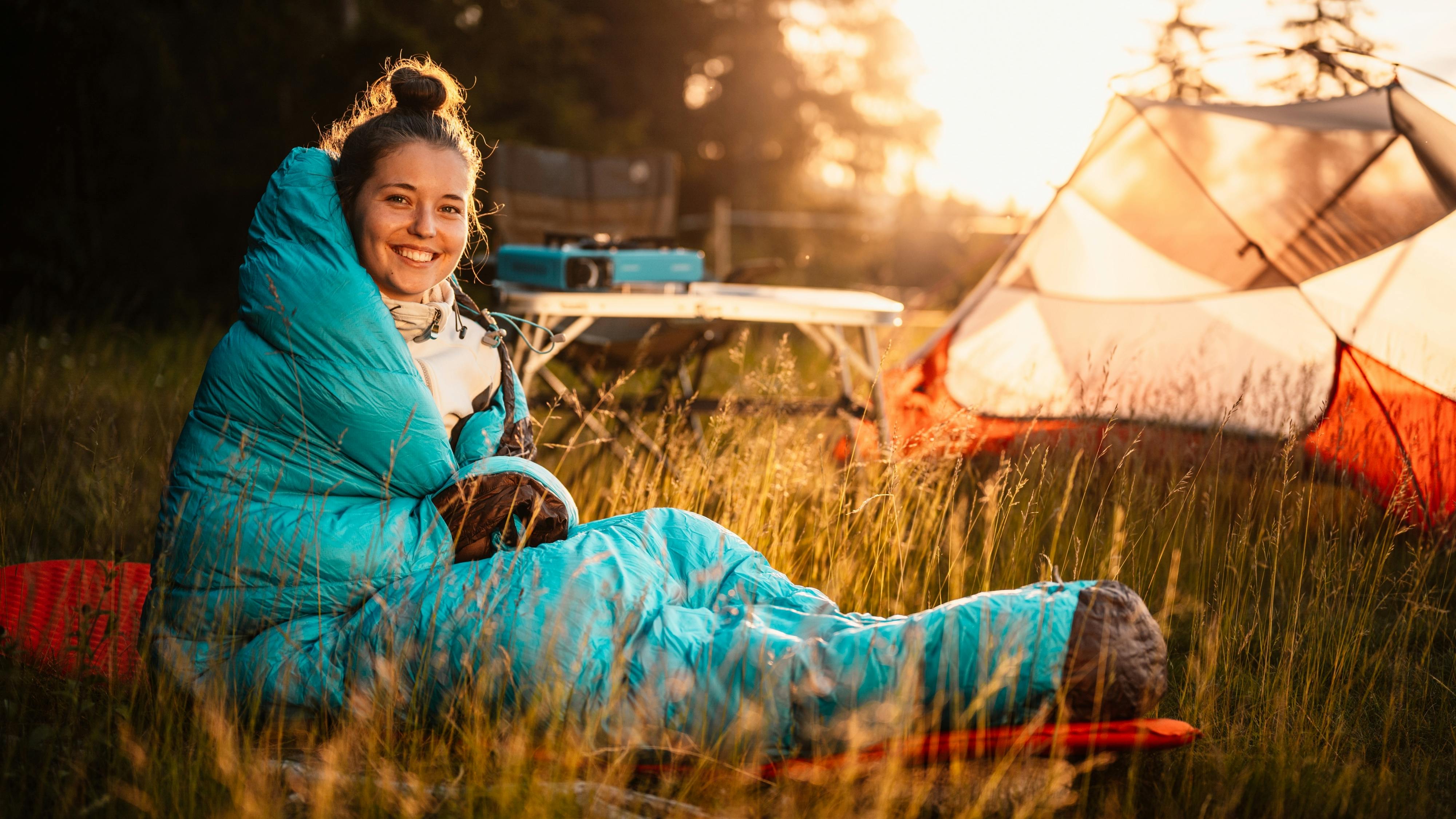 A woman in a blue sleeping bag sits outside her tent and smiles at the camera. 