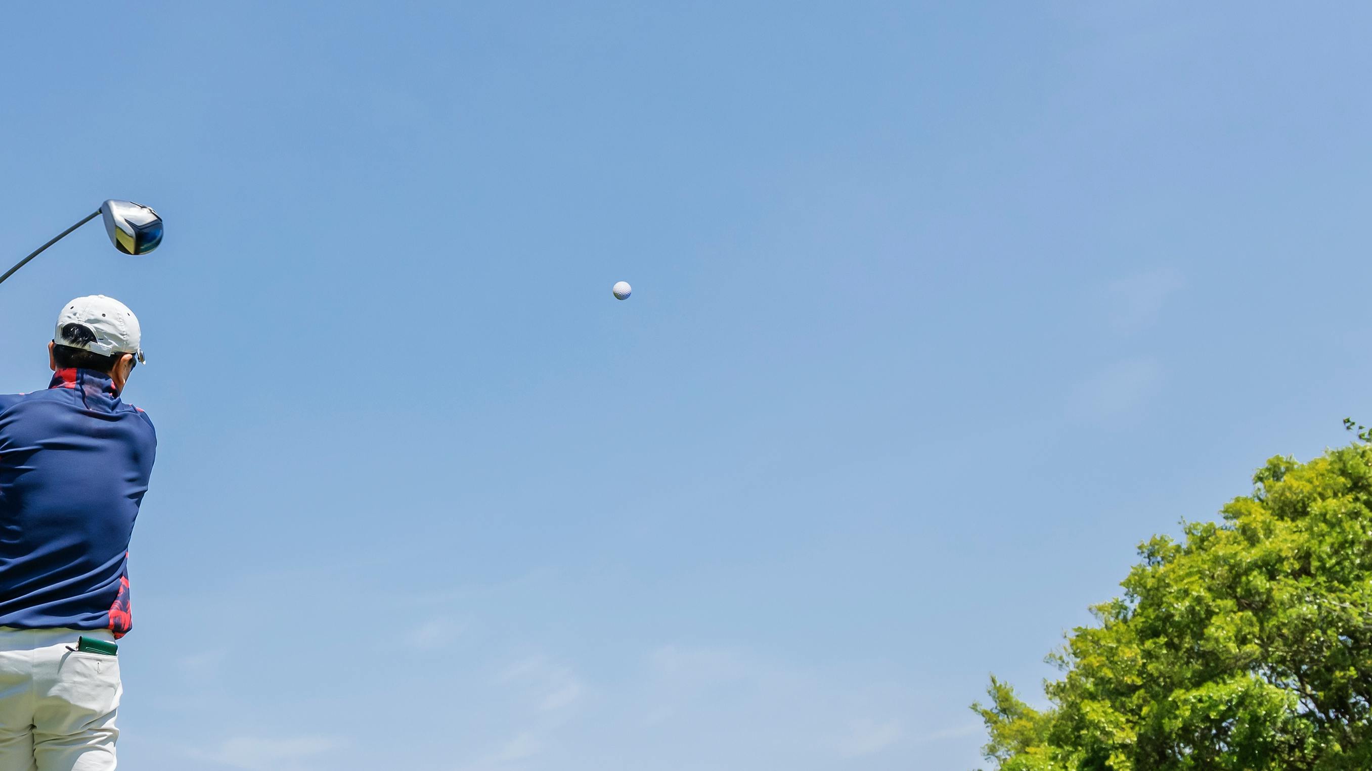 A golfer swinging his driver as a golf ball flies through the air.