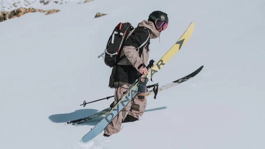 A skier holding his Dynastar skis as he walks through the snow.