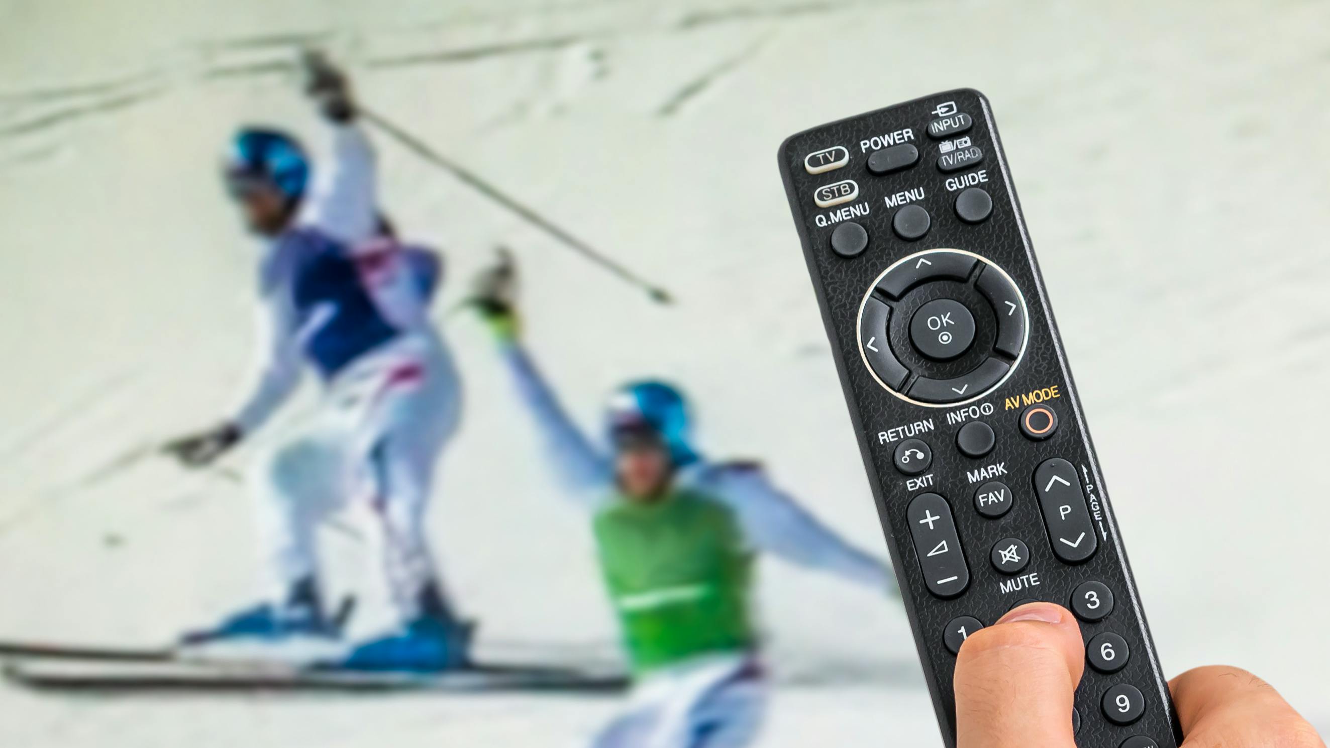 Close up of a remote control with a television showing some skiers in the background. 