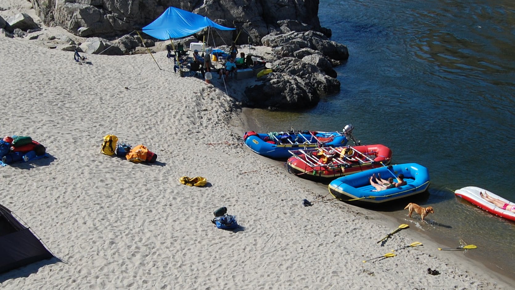 Three kayaks on the shore of a river. There is some gear and two tents set up on the shore. 