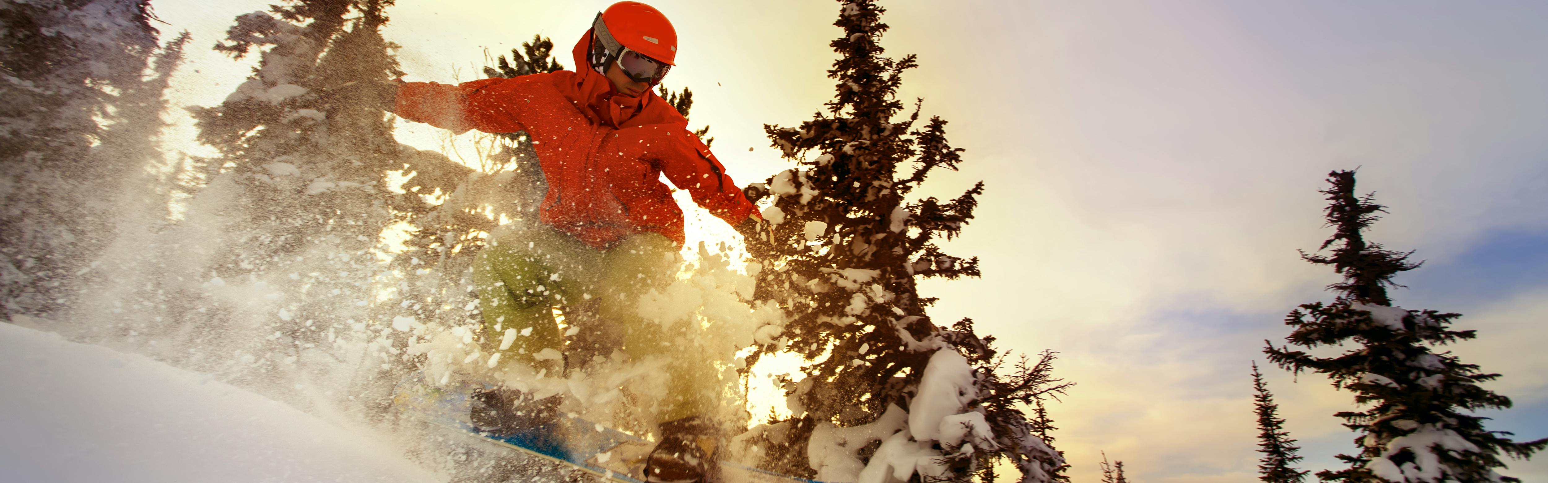 A man snowboarding down a powdery trail. There are trees and a sunset behind him. 