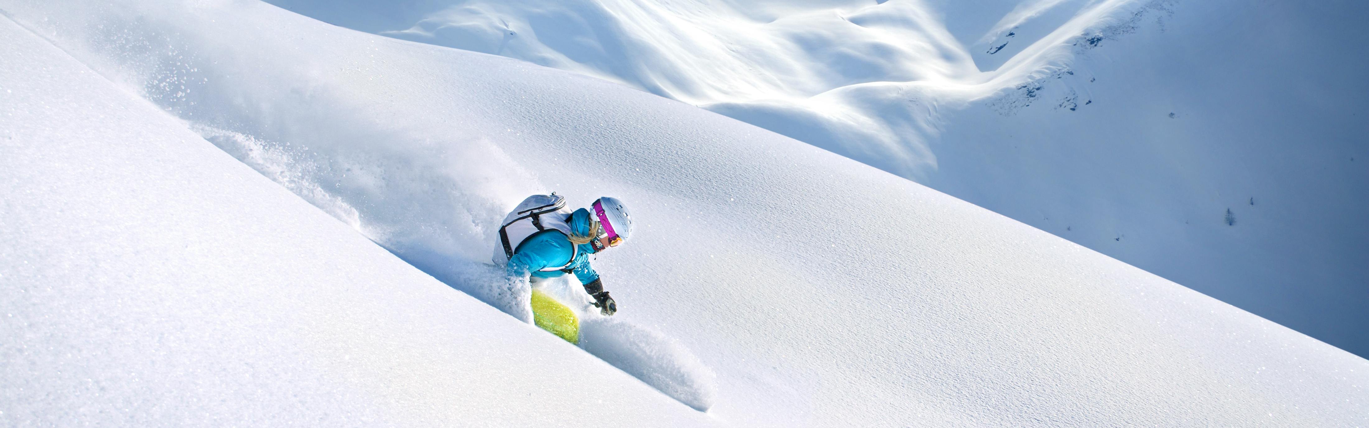 A skier turns down a snowy slope with her white backpack, white helmet, and blue jacket. 