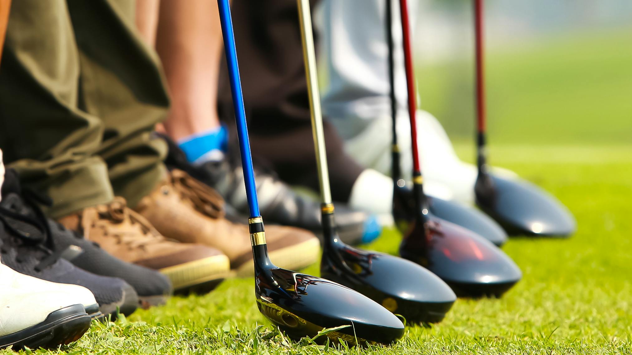 Several golfers standing in a row holding their golf clubs. Only their shoes and the shaft of the golf clubs are visible. 
