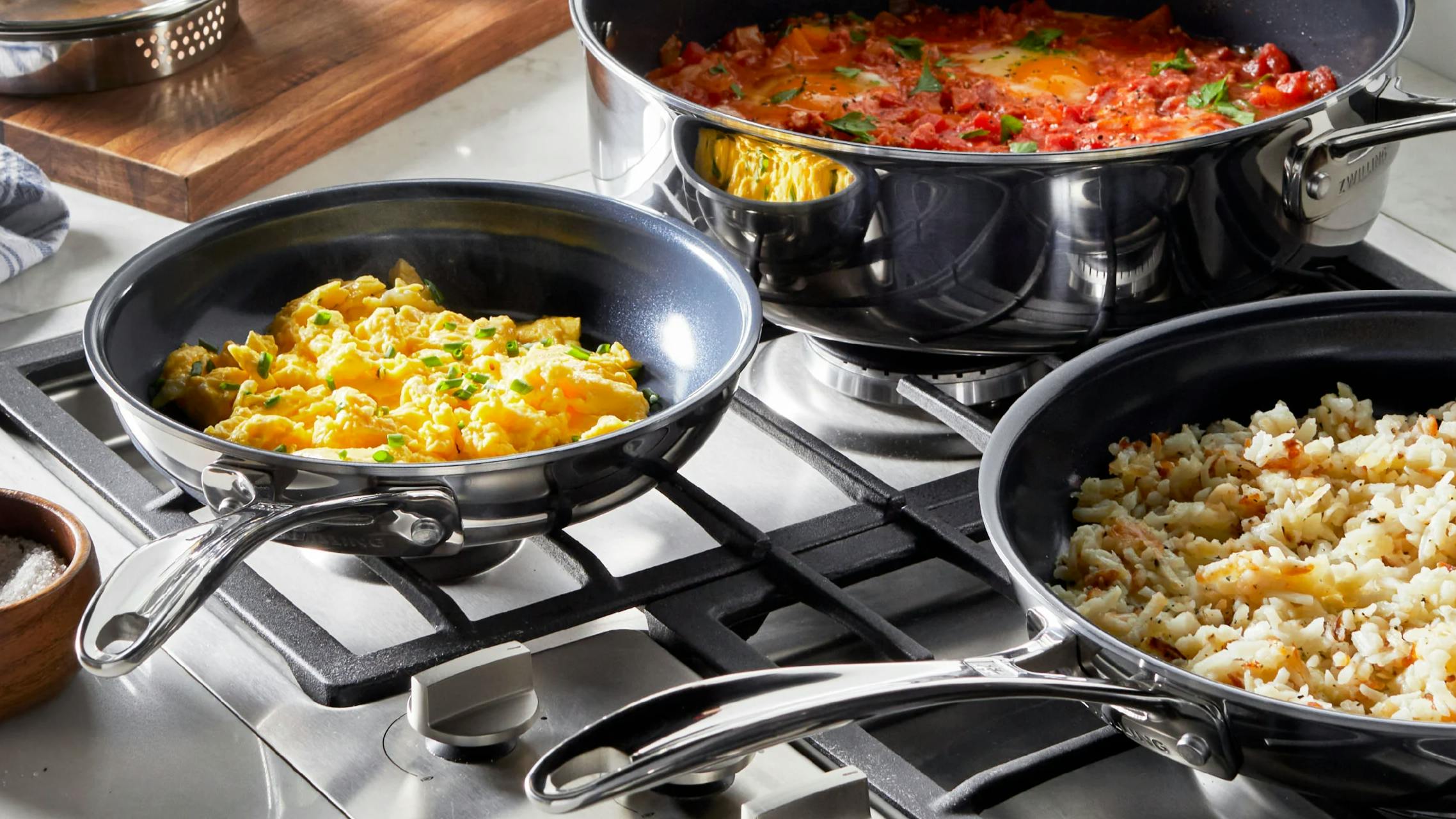 The Zwilling Clad CFX Stainless Steel Ceramic Nonstick Cookware Set · 10-Piece Set. The pans are on the stove with food in them. 