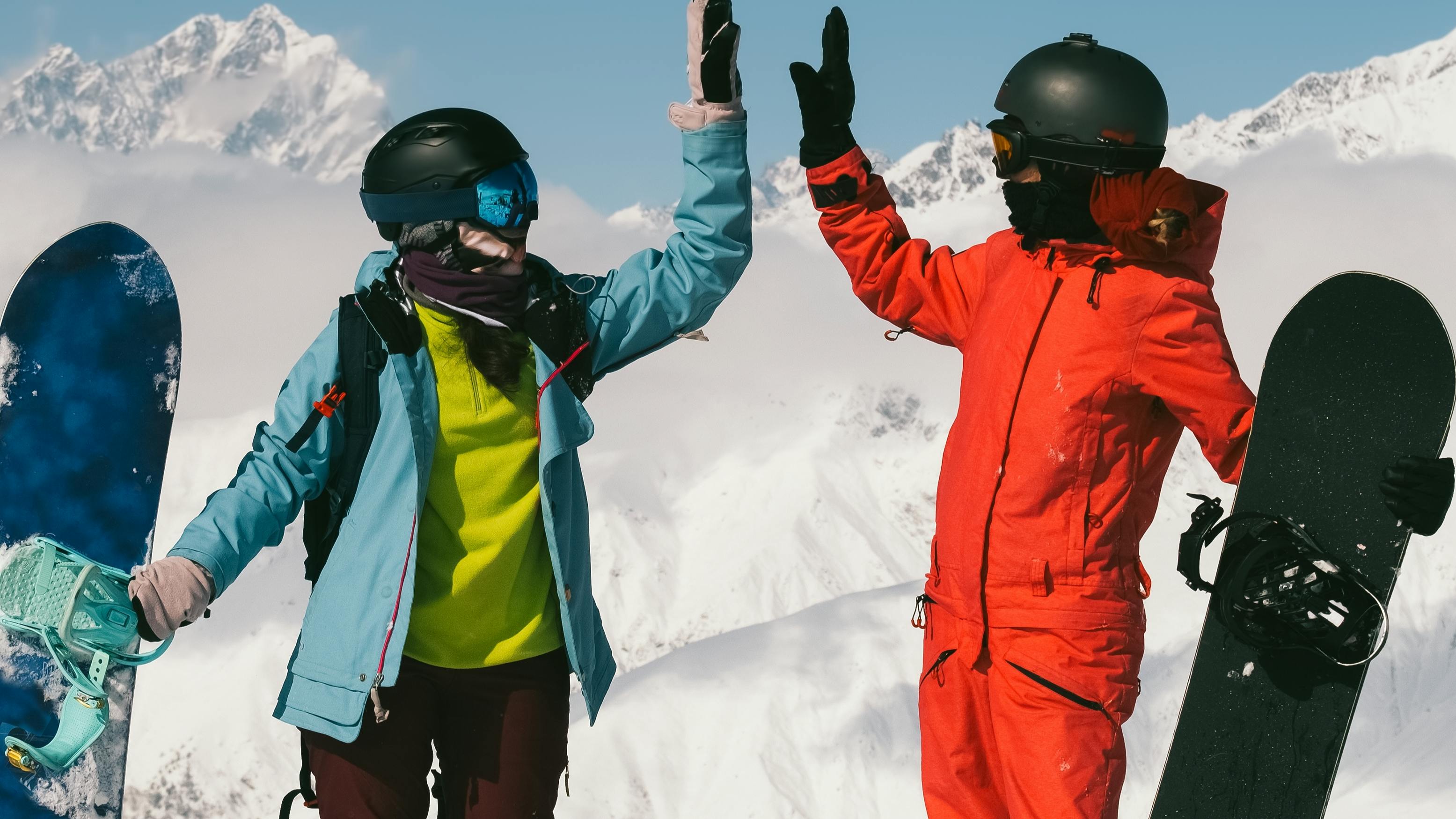 Two snowboarders standing at the top of a mountain as they high-five. They are both wearing full snowboard gear and holding their snowboards. 