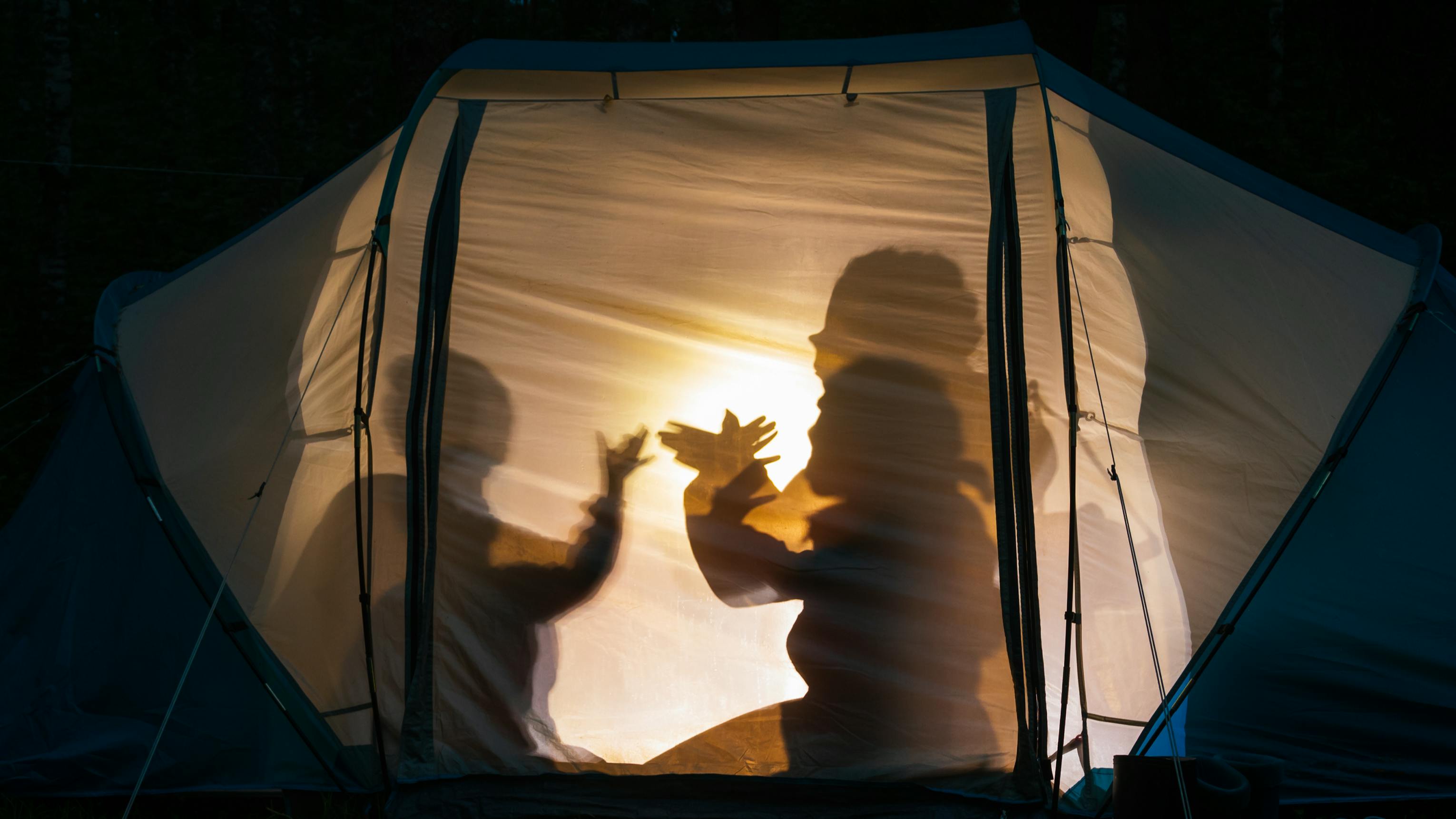 Two campers in a tent making shadow puppets with a light behind them. 