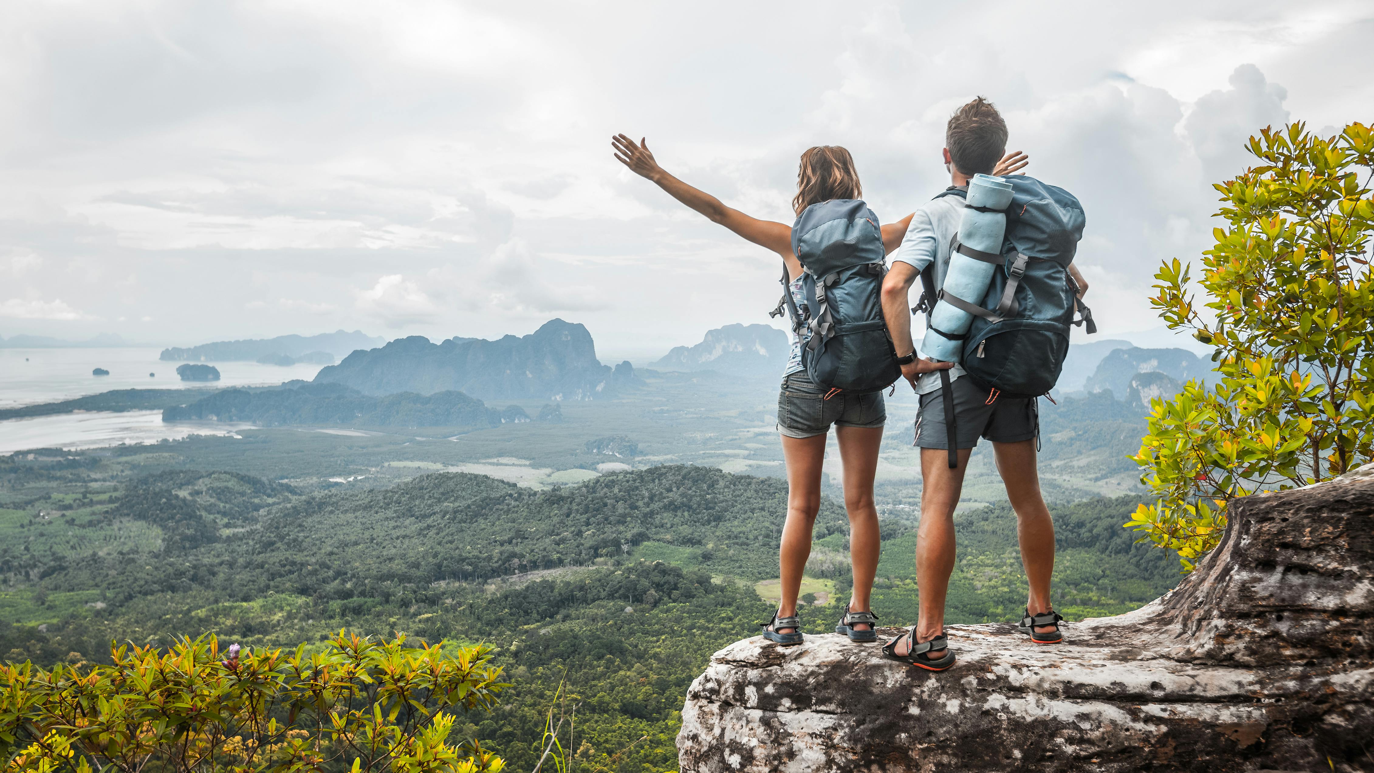 Two backpackers stand at the top of an outlook as they are both wearing their backpacking bags. The woman has her arms outstretched. 