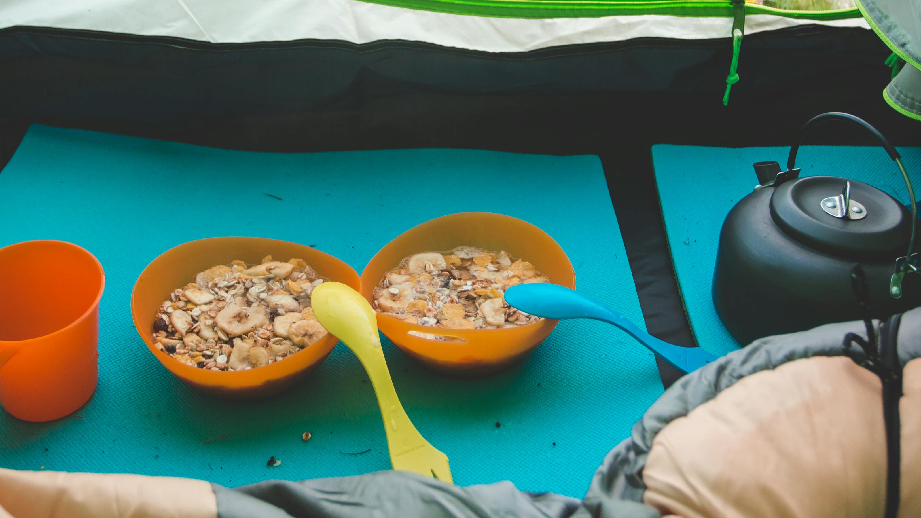 Two bowls of oatmeal in plastic bowls. They each have a camping spoon in them and there is a plastic cup sitting next to the bowls. You can also see a kettle. The kettle, bowls, and cup are on a sleeping pad inside a tent. 