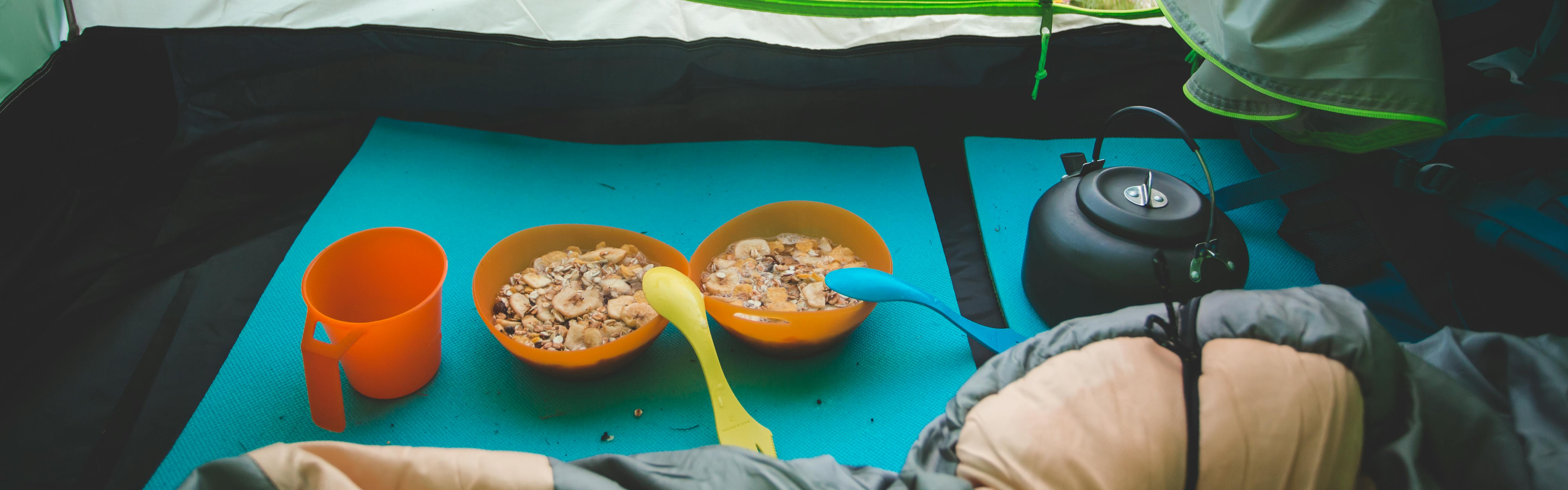 Two bowls of oatmeal in plastic bowls. They each have a camping spoon in them and there is a plastic cup sitting next to the bowls. You can also see a kettle. The kettle, bowls, and cup are on a sleeping pad inside a tent. 