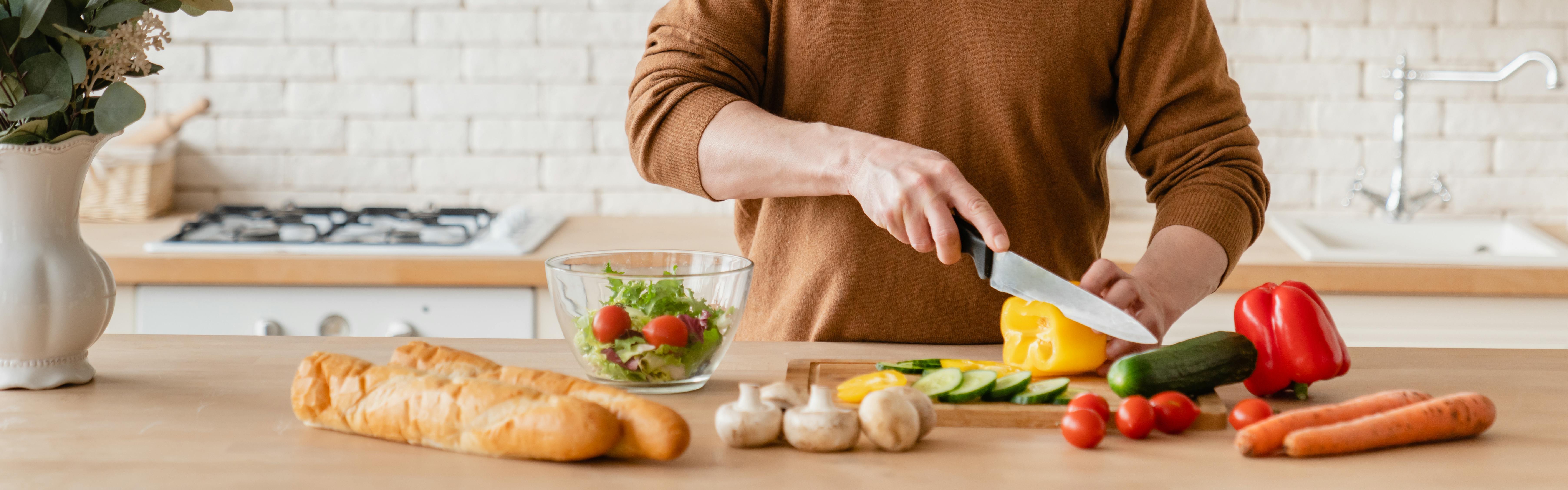 Close up of some hands chopping a bell pepper on a counter with a bowl of salad and some bread laying next to it. 