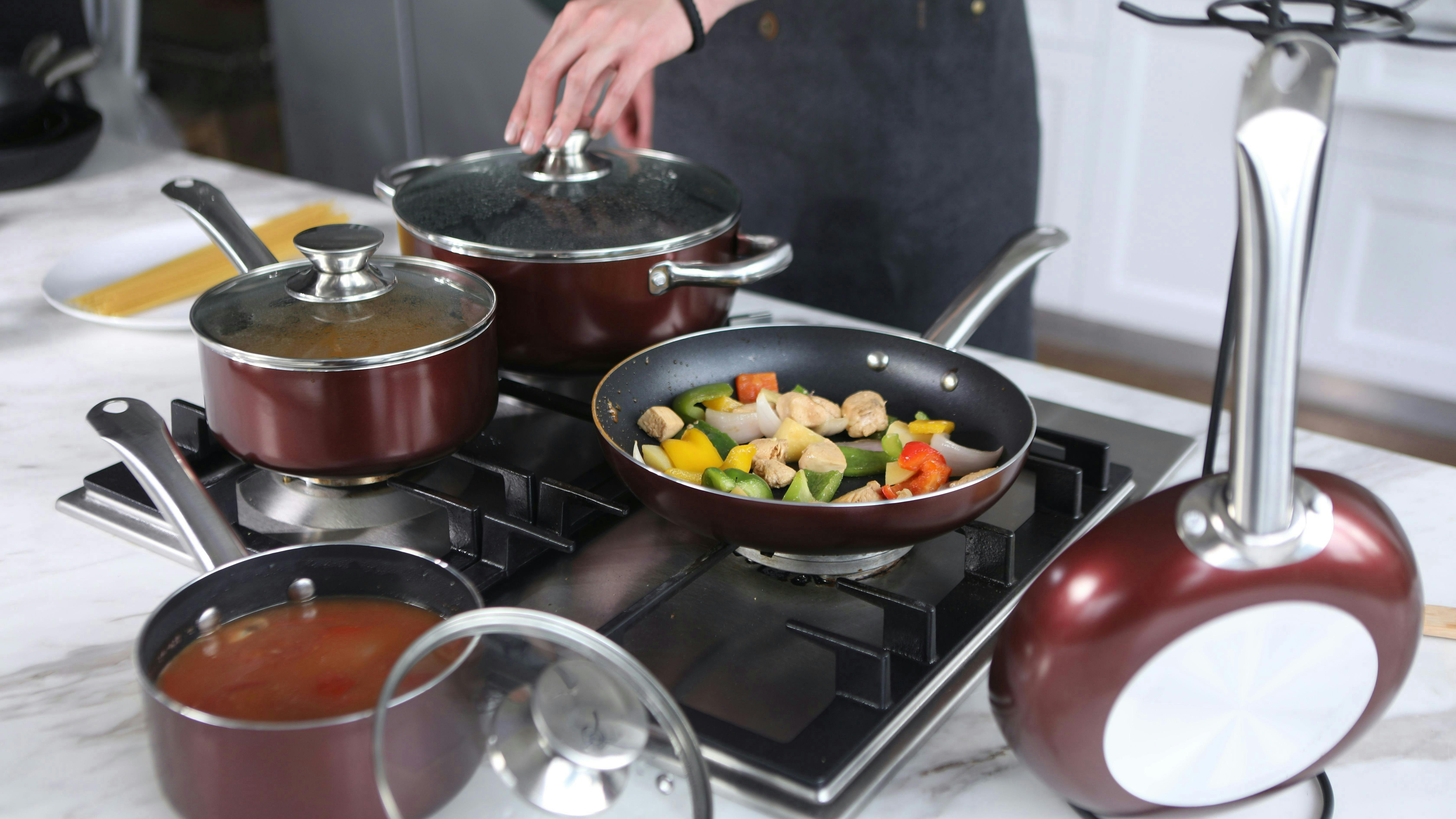 Cookware on stovetop. One pot has soup in it and one pan has vegetables in it. There is a hand taking the lid off of one of the large pots still on the stove. 