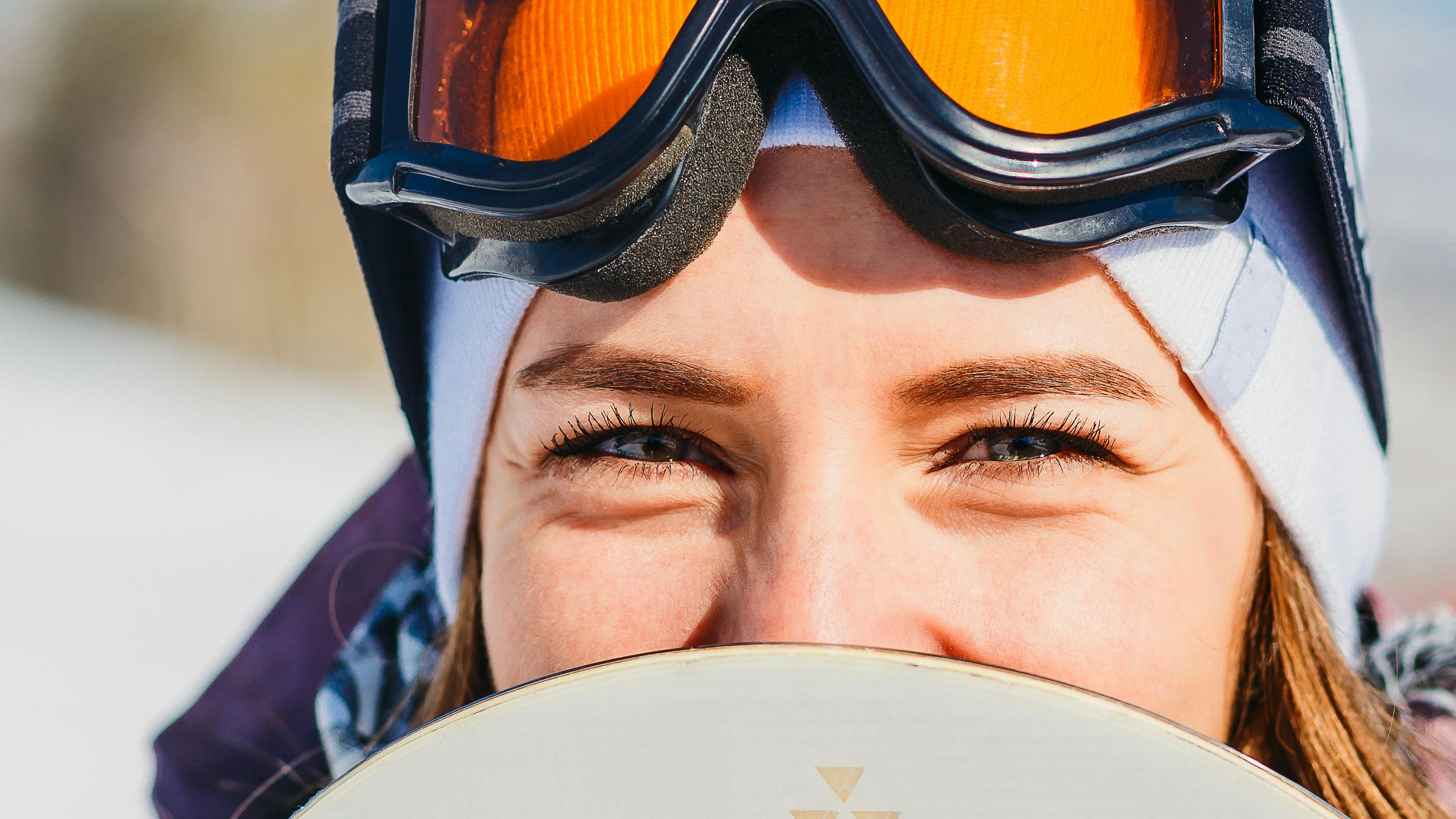A snowboarder with goggles on her head smiling behind her snowboard.