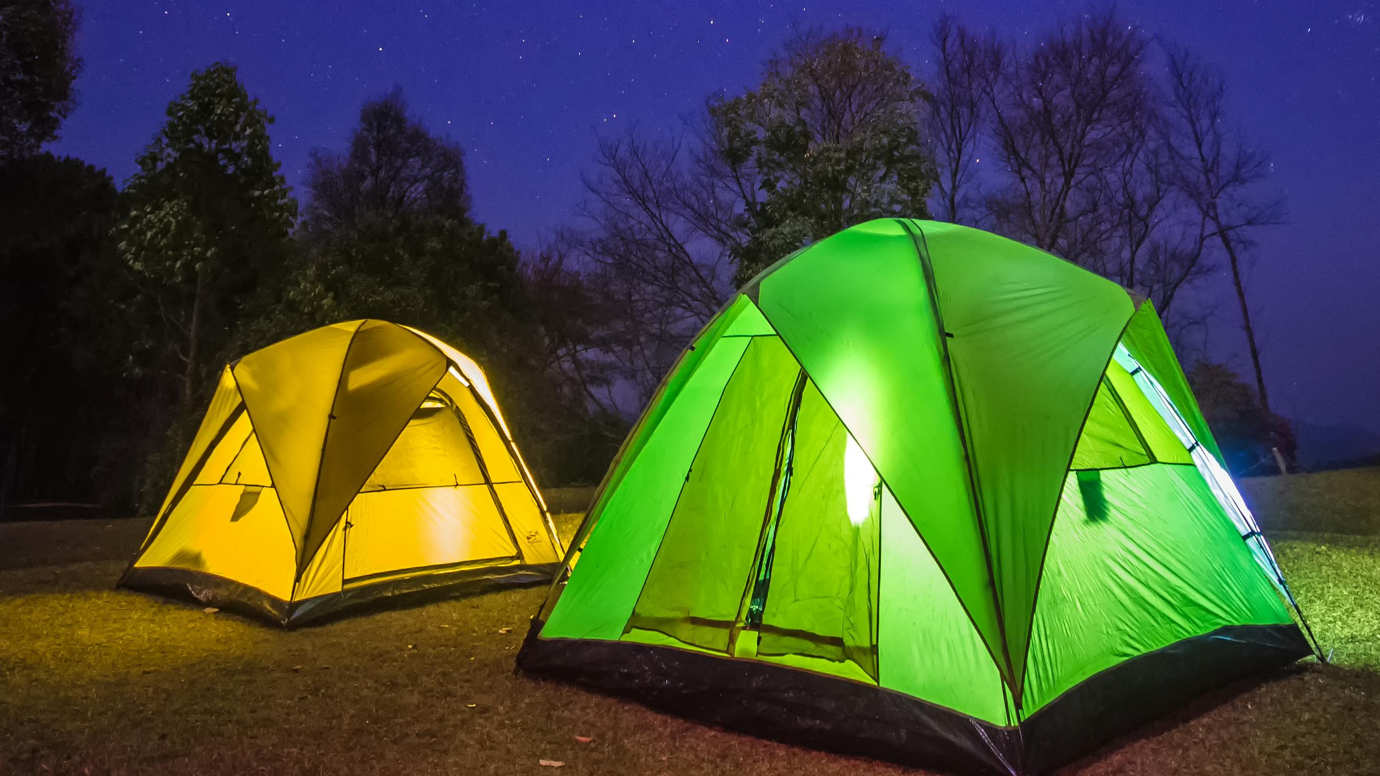 Two tents set up on a dark night. One is green and one is yellow and they are lit up from the inside. 