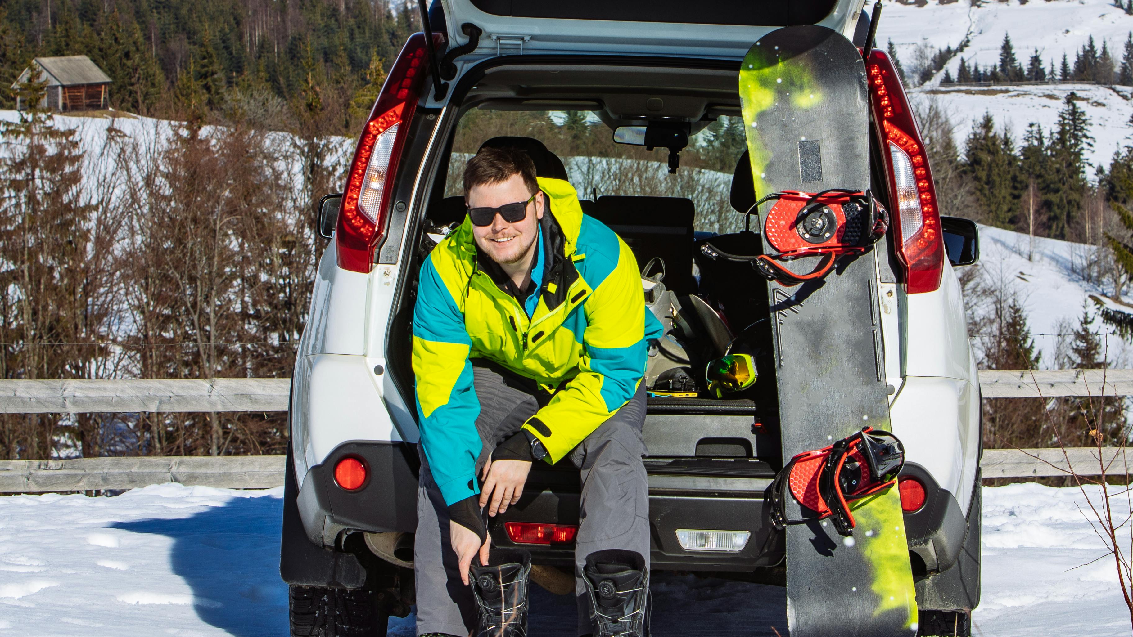 A man is sitting in the back of his car as he smiles at the camera. There is a snowboard sitting in the back of his car with him and he is putting snowboard boots on. The parking lot is snowy and there are snowy hills behind him. 