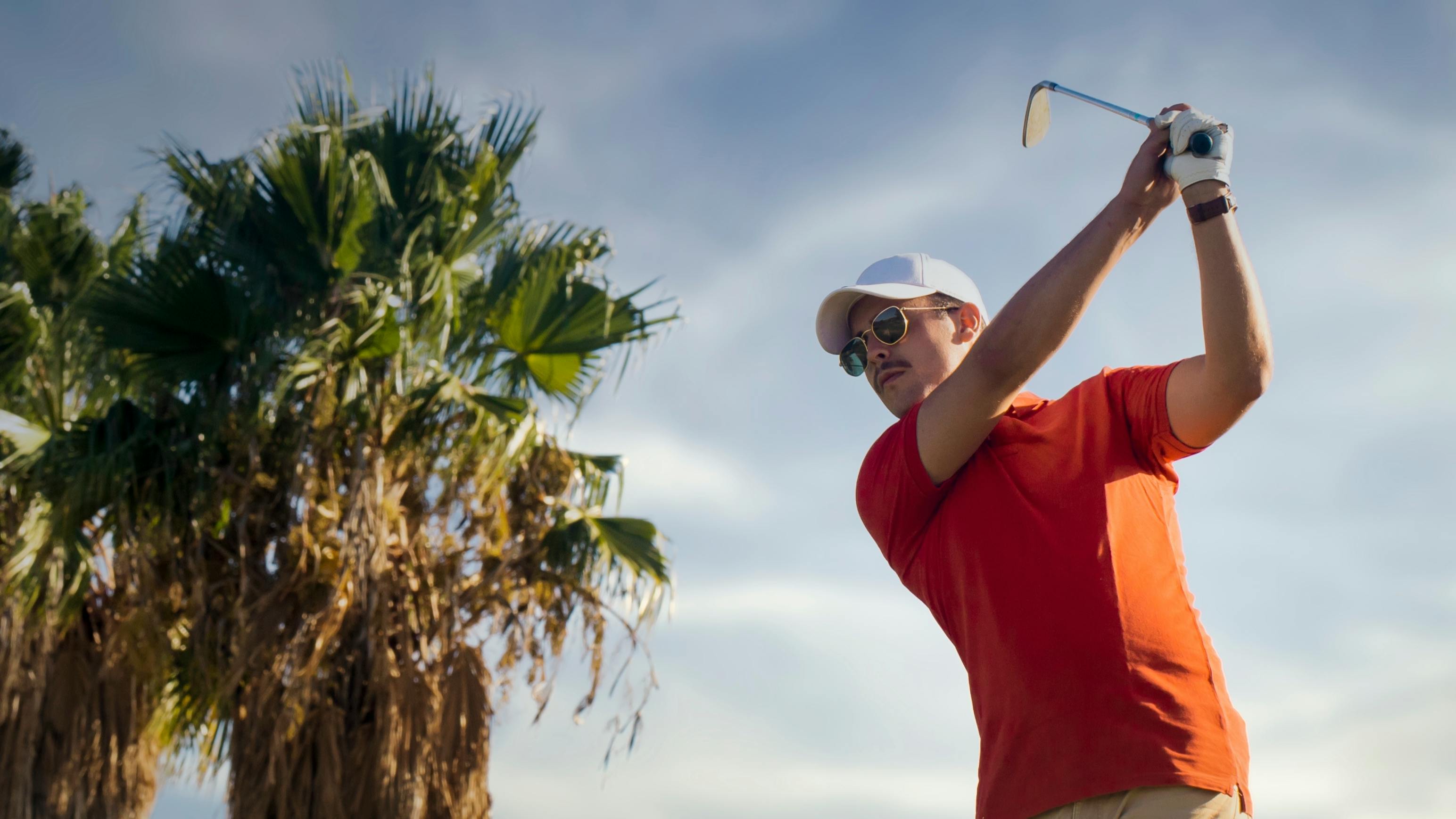 A golfer on his backswing. He is using an iron and there is a palm tree in the background. 
