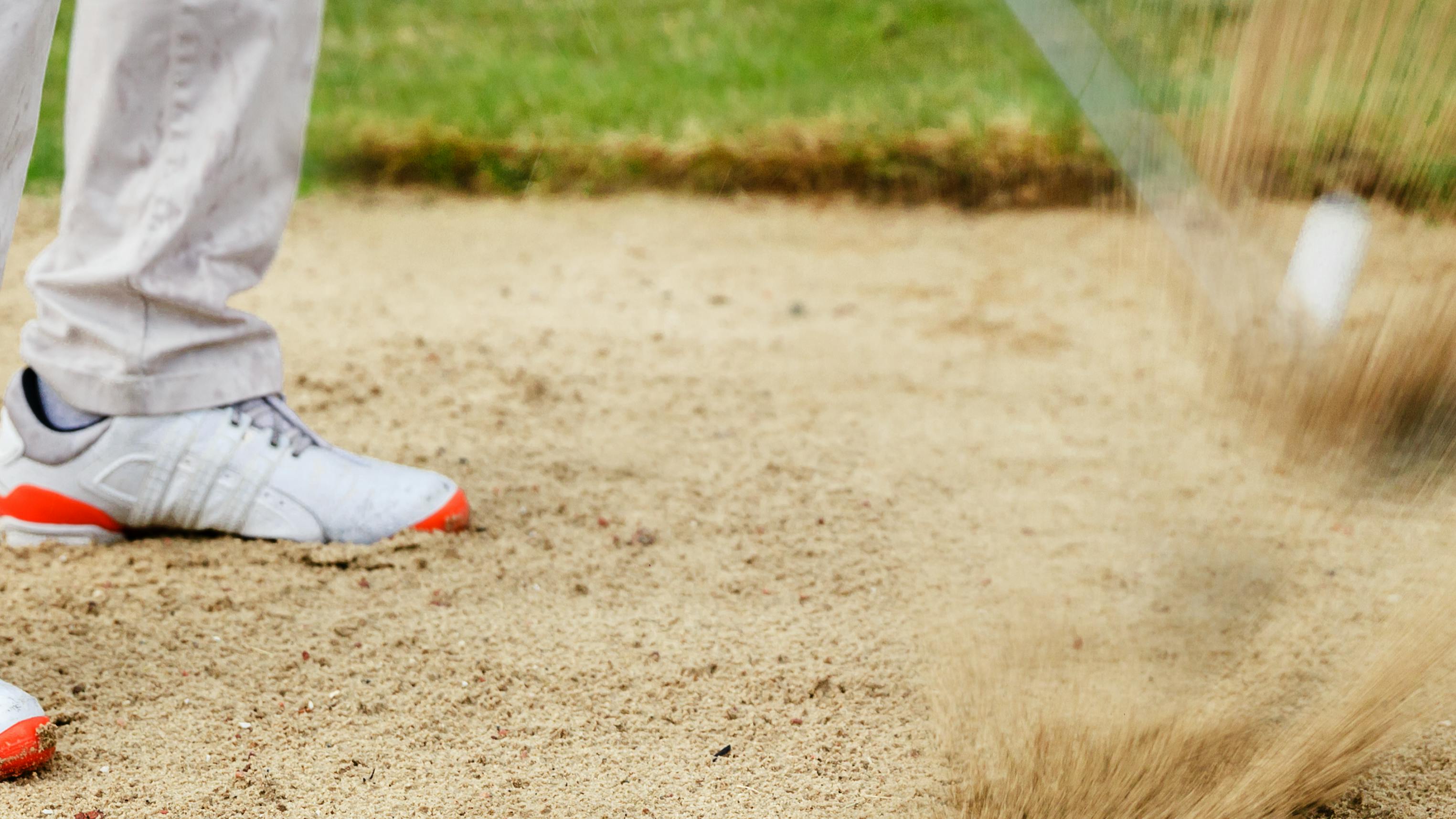 A golfer with rain droplets on his pants hits a golf ball out of a bunker. 