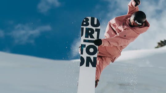 A snowboarder doing a trick as the bottom of her Burton snowboard is showing. 