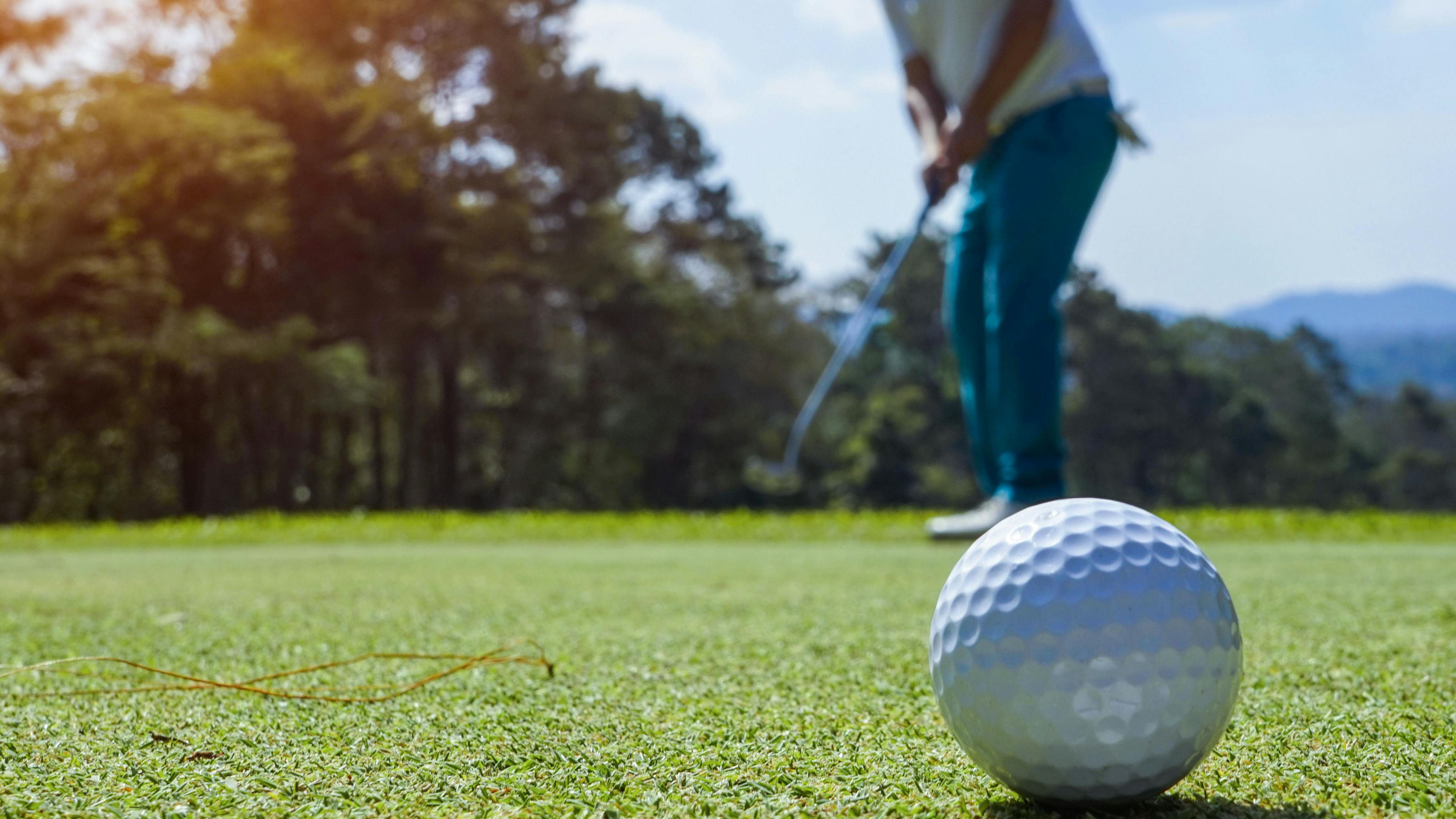 Close up of a golf ball rolling as a golfer stands in the background holding a putter.