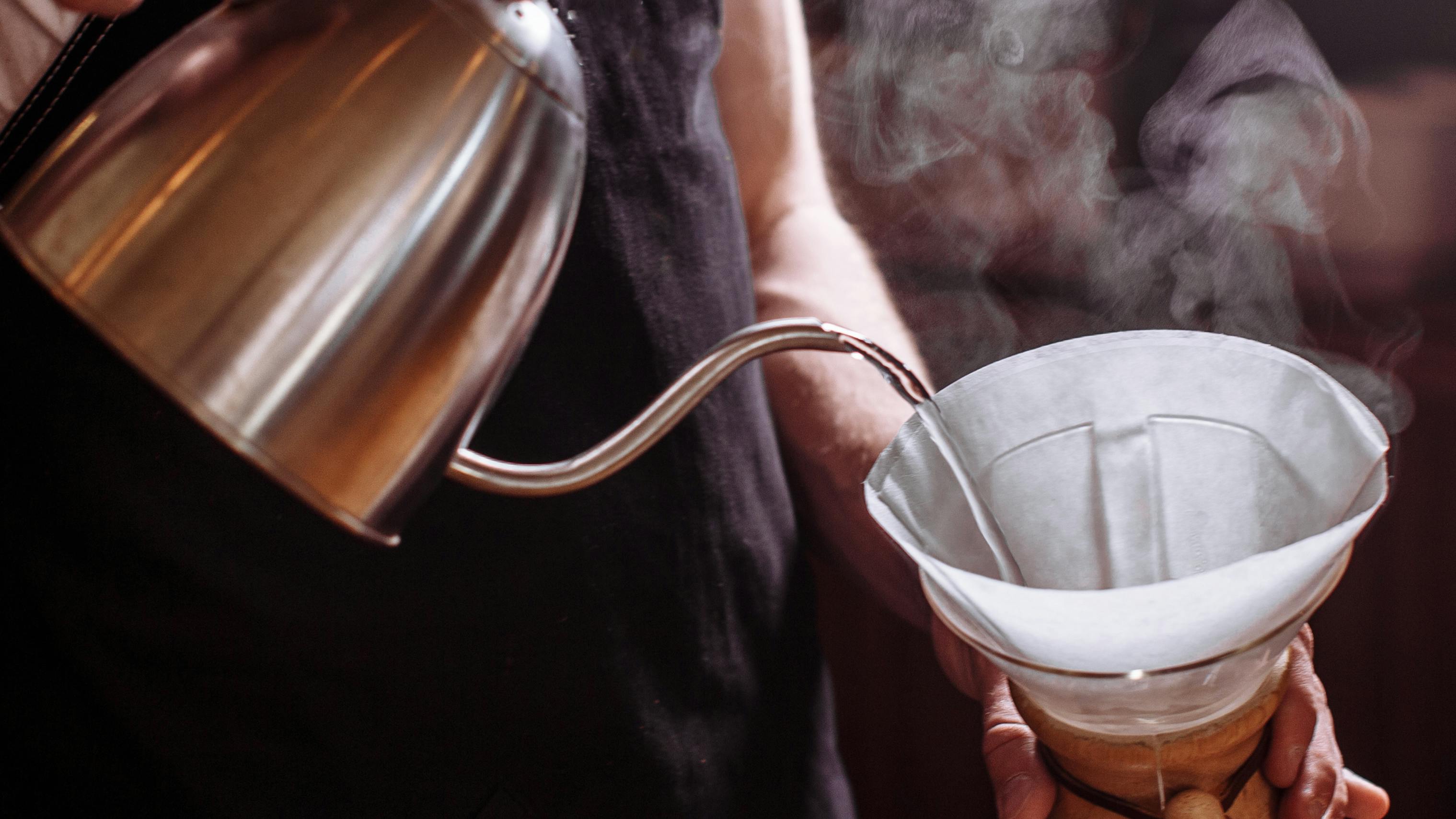 A person pouring water in a pour over coffee maker from a gooseneck pitcher. 