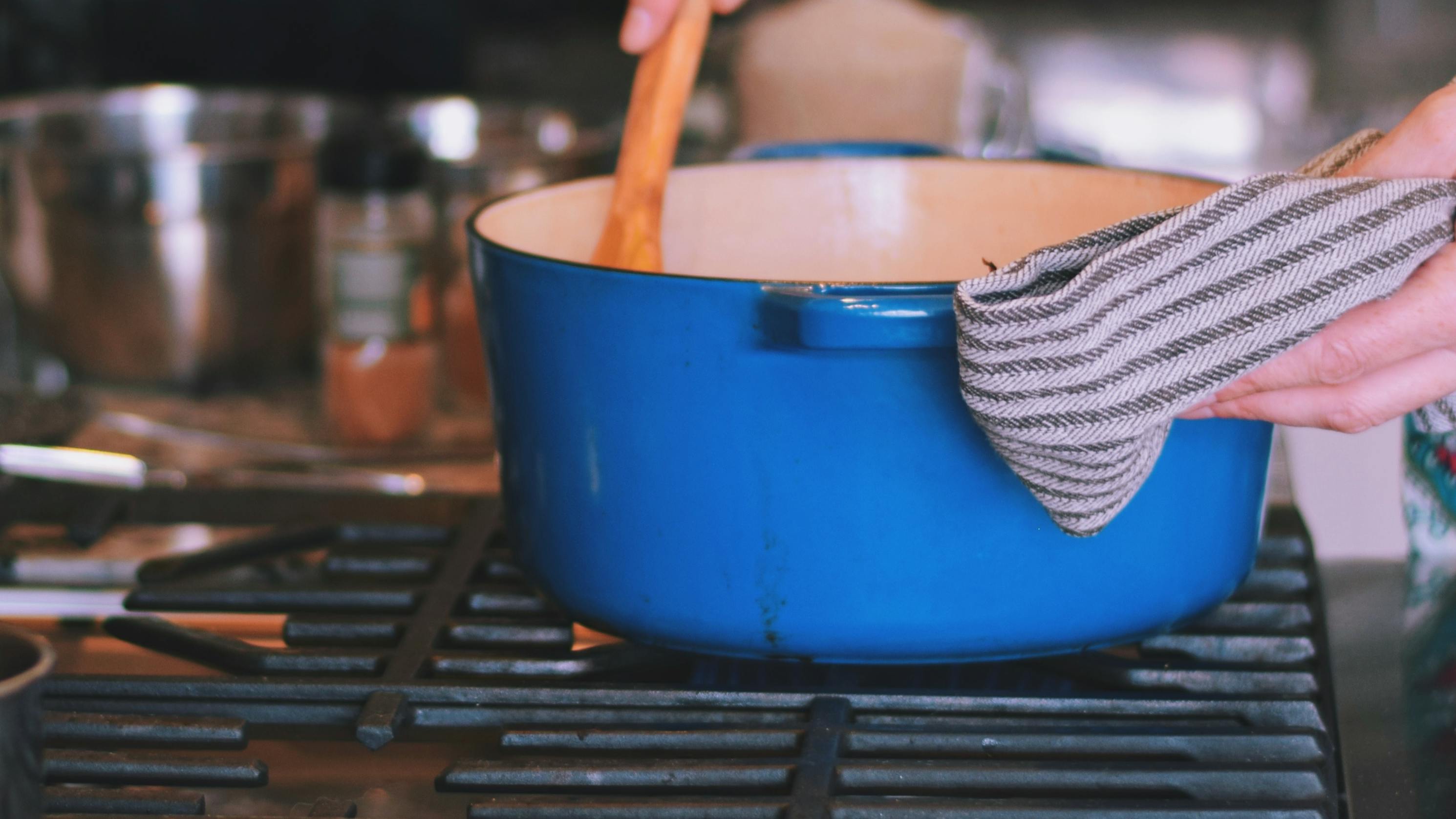 A blue cast iron dutch oven on a stove as a woman stirs the contents with a wooden spoon. 