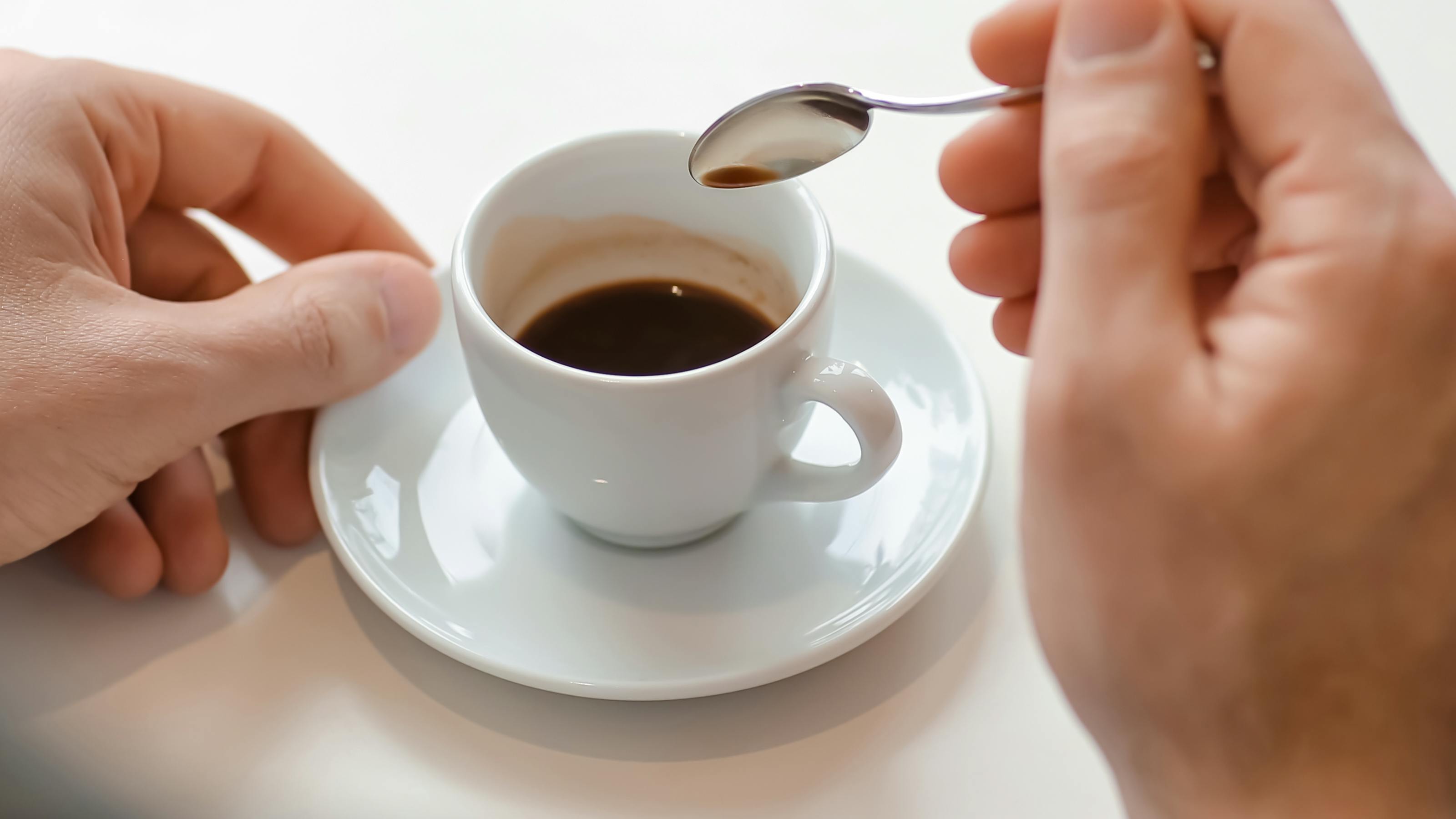 A cup of espresso on a white plate with a person holding a spoon over it. 