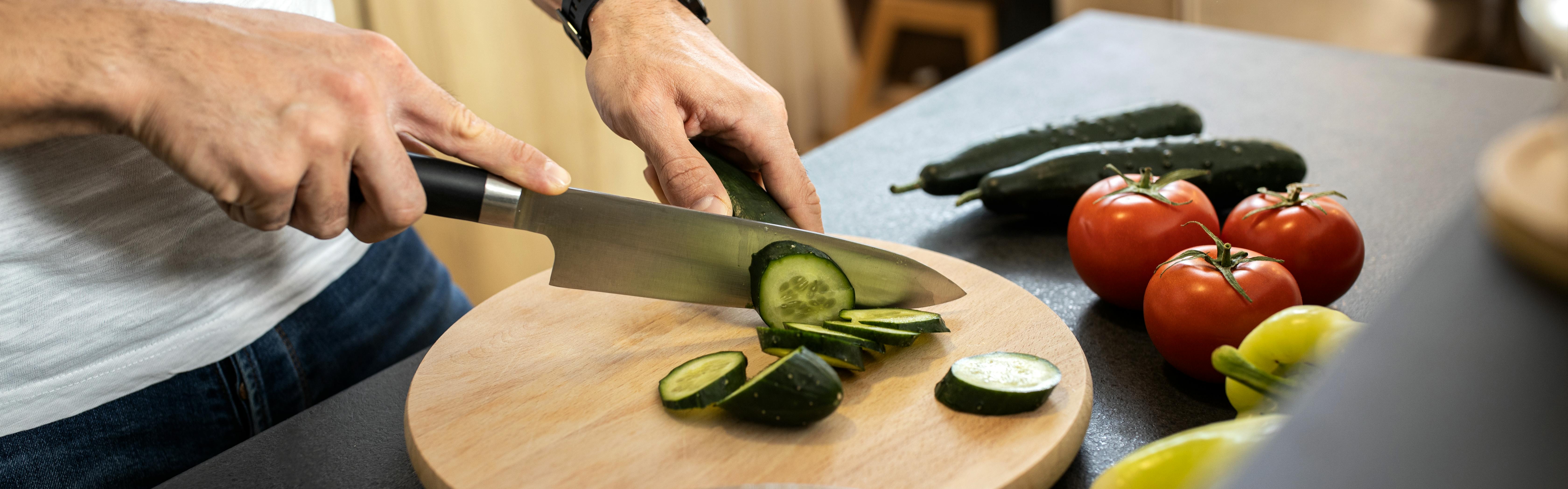 A knife chopping a cucumber on a wooden cutting board. 