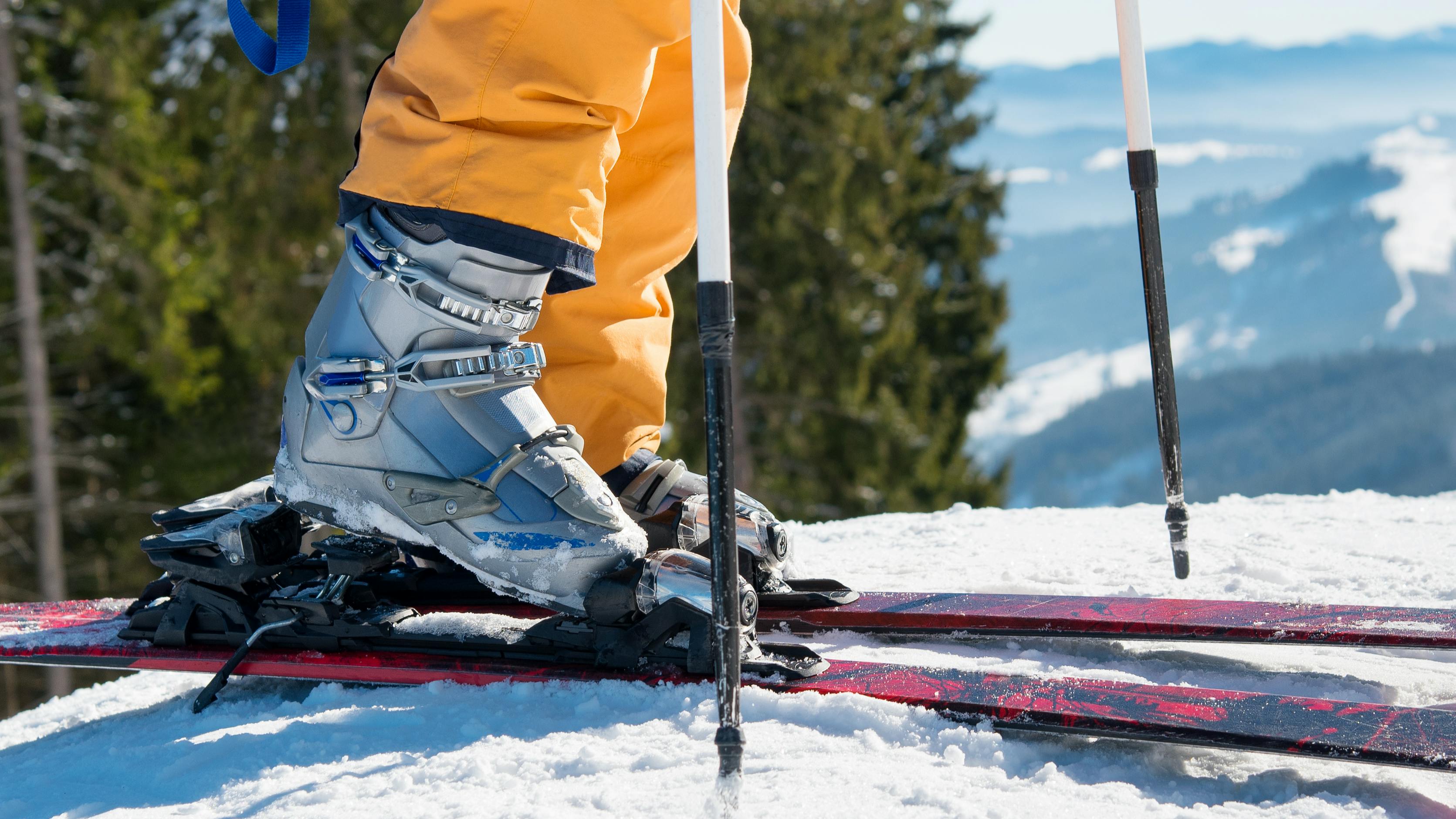 A skier clicking their boot into their ski bindings as they stand on top of a snowy mountain. 